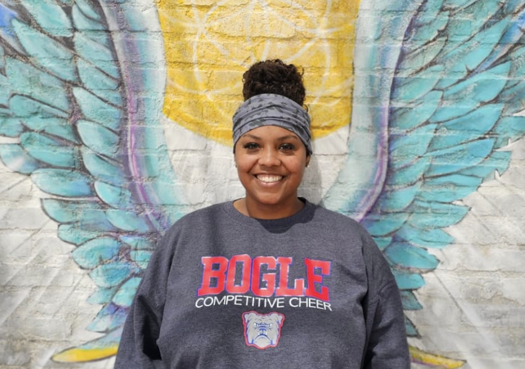 A woman smiling, wearing a gray headband and a gray sweatshirt that says "BOGLE Competitive Cheer" with a bulldog mascot, standing in front of a mural of angel wings painted on a brick wall.