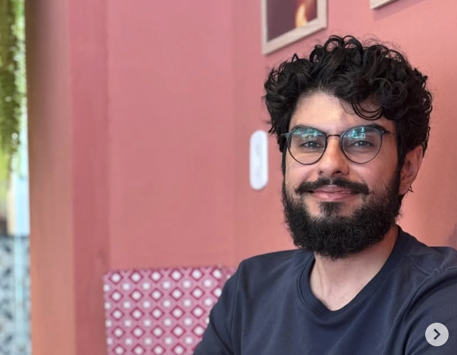 A man with curly black hair, glasses, and a beard smiling at the camera, sitting in a cafe with pink walls and framed pictures behind him.