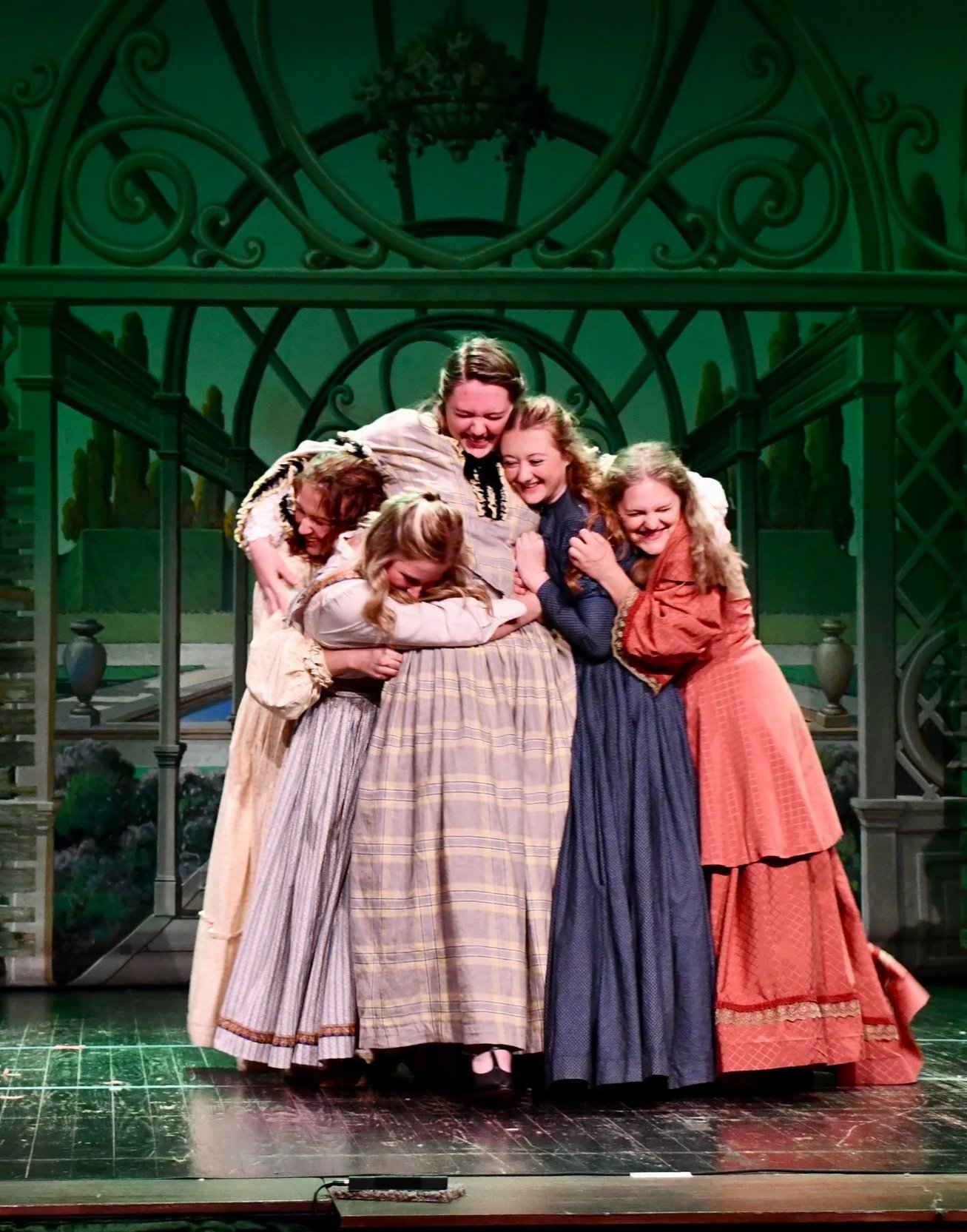 The cast of a theatrical play on stage applause and hug, with a backdrop of a garden or greenhouse scene.