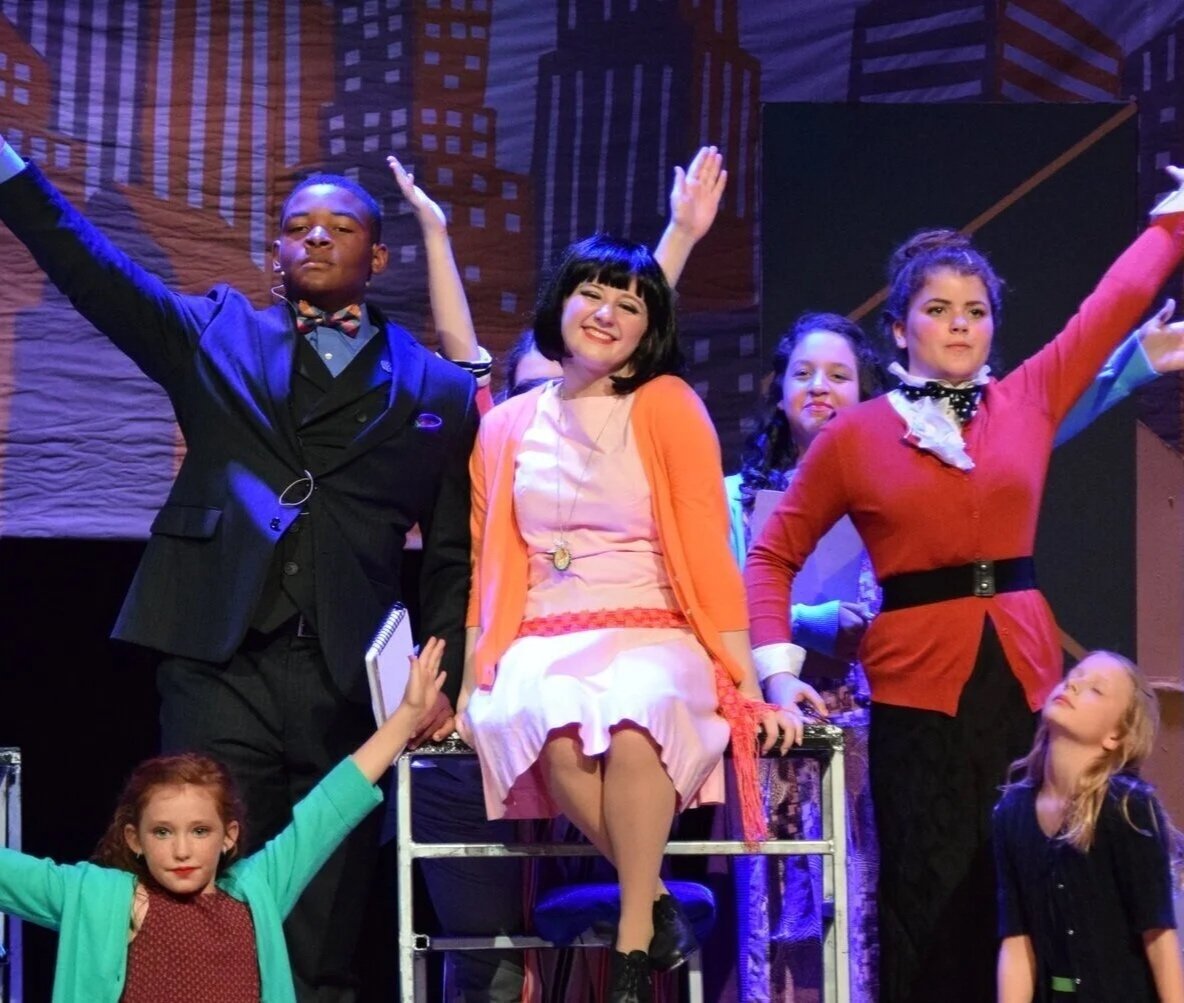 A group of children and teenagers performing on stage, dressed in colorful costumes, with some holding clipboards and notebooks, against a backdrop of city buildings.