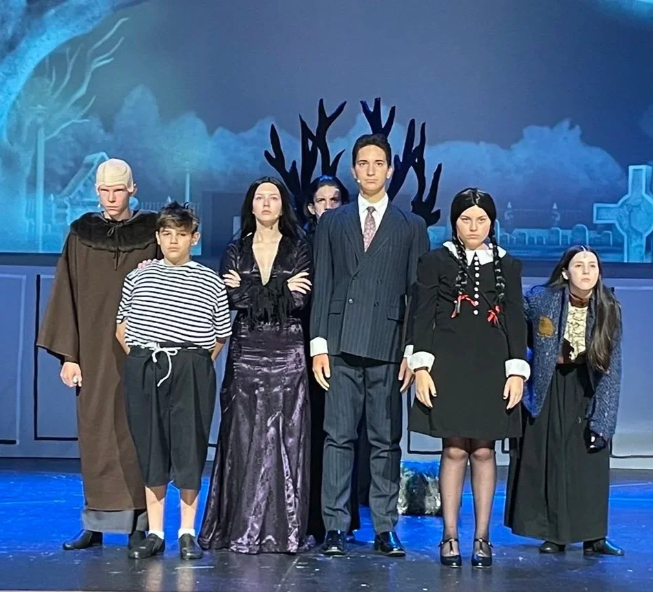 A group of six people in gothic costumes on stage, with a dark, eerie background featuring large black antlers and tombstones.