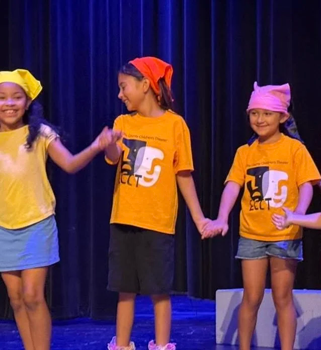 Four young girls holding hands on stage during a performance. Two girls wear yellow shirts, one with a pink headscarf, and the other with a blue headscarf. Two girls wear orange shirts, one with a red headscarf and the other with a pink headscarf. Th