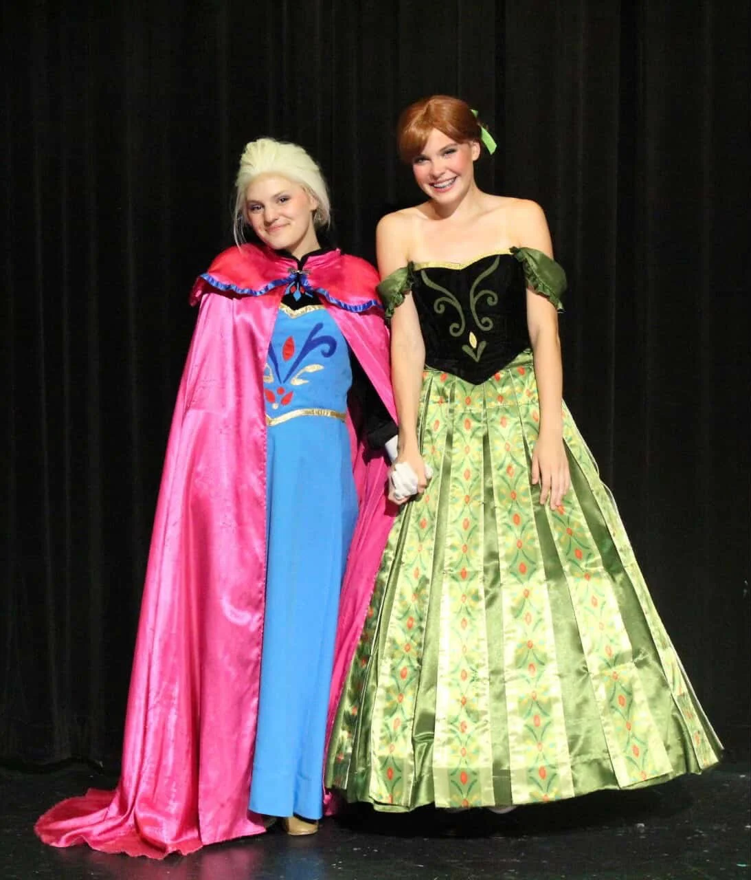 Two women dressed as princesses holding hands and smiling on a stage with black curtains in the background.