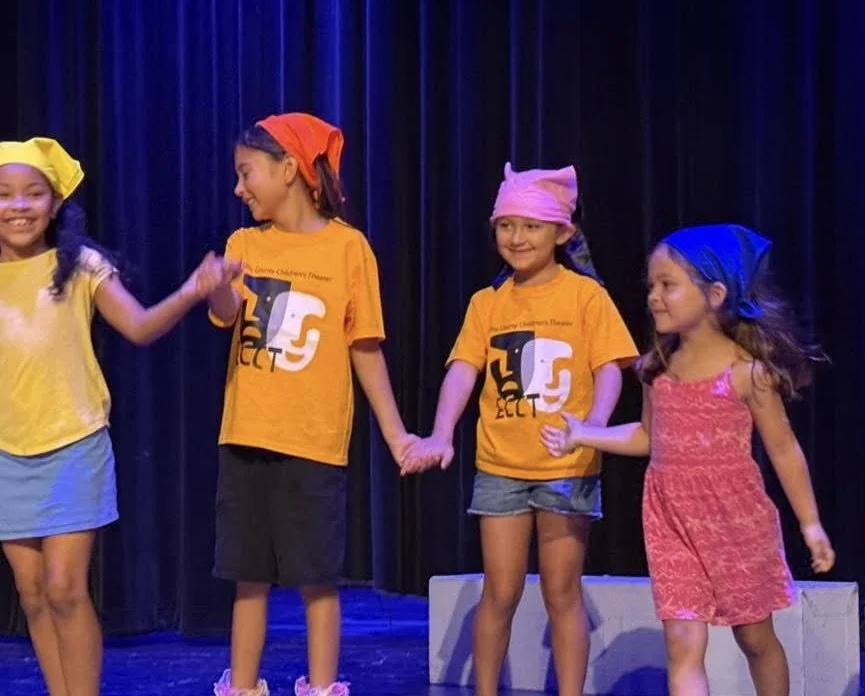 Four young girls holding hands on stage during a performance. Two girls wear yellow shirts, one with a pink headscarf, and the other with a blue headscarf. Two girls wear orange shirts, one with a red headscarf and the other with a pink headscarf. Th