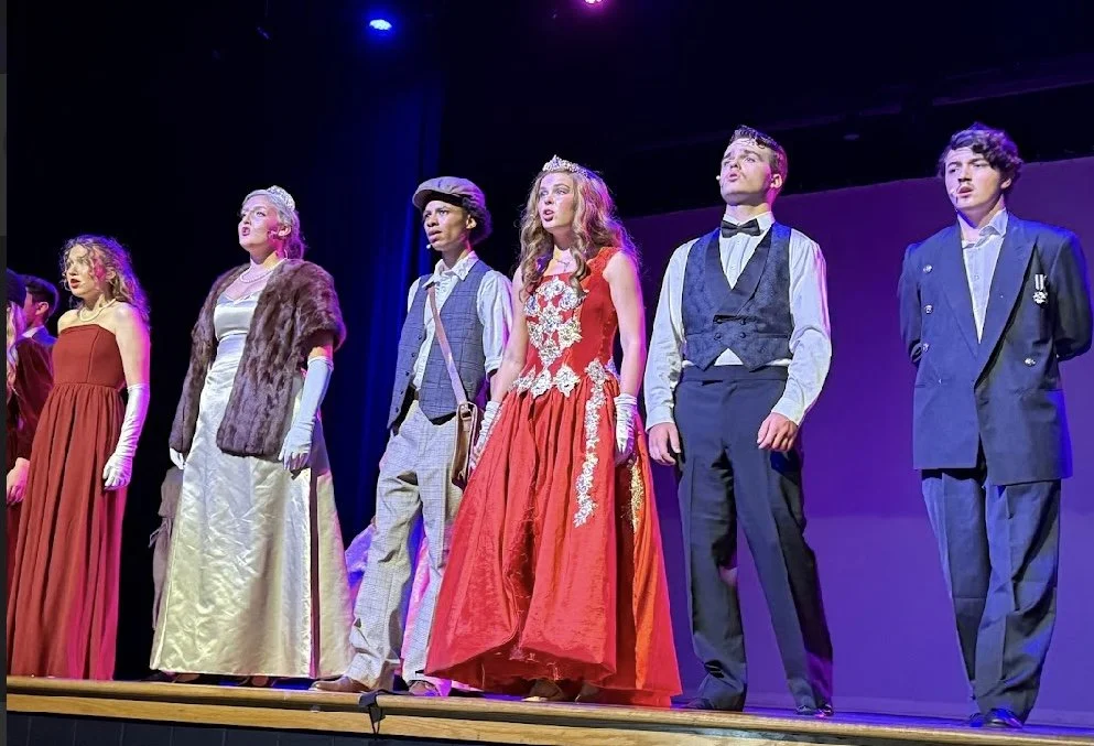 Theater cast on stage in period costumes, performing in a musical or play, illuminated by stage lighting.