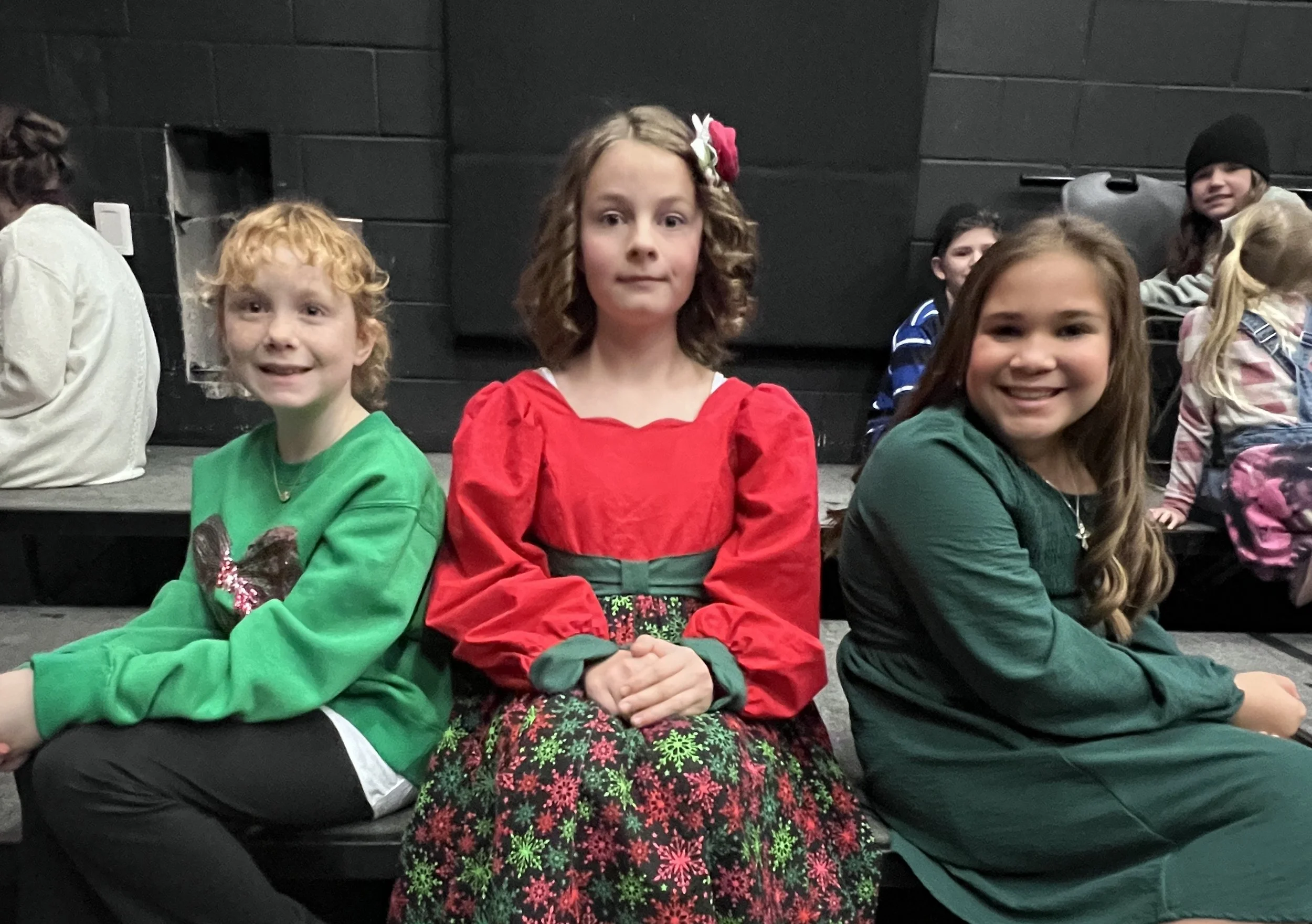 Three young girls sitting on black seats in a theater or auditorium, dressed in festive clothing, smiling at the camera.