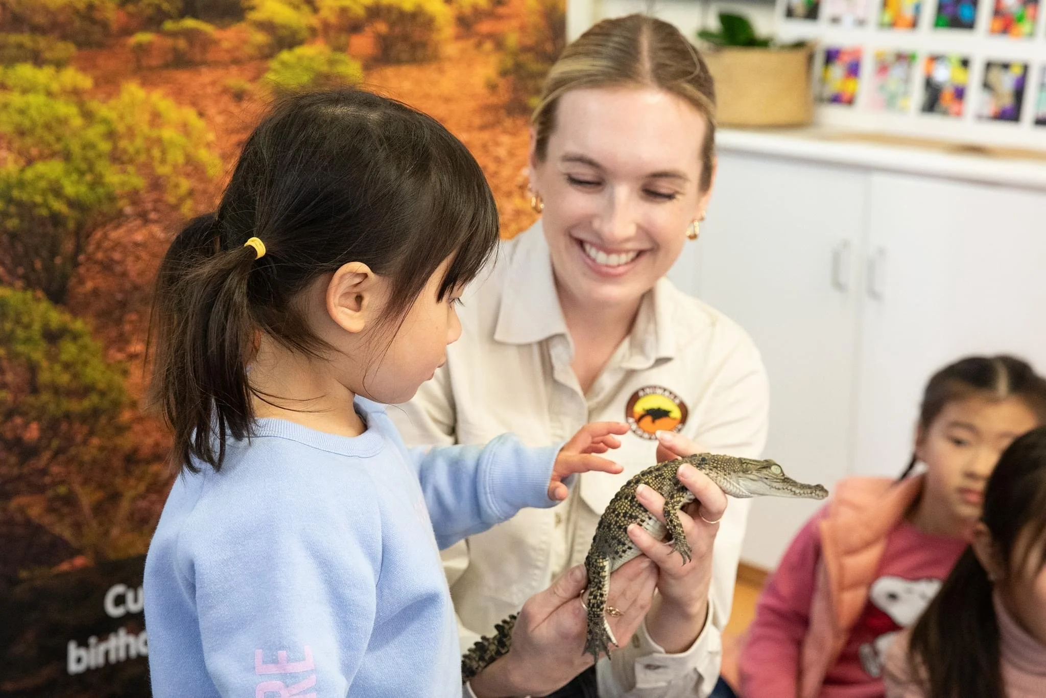 A woman holding a baby freshwater crocodile and smiling at a young girl who is reaching out to touch it during an educational wildlife presentation.