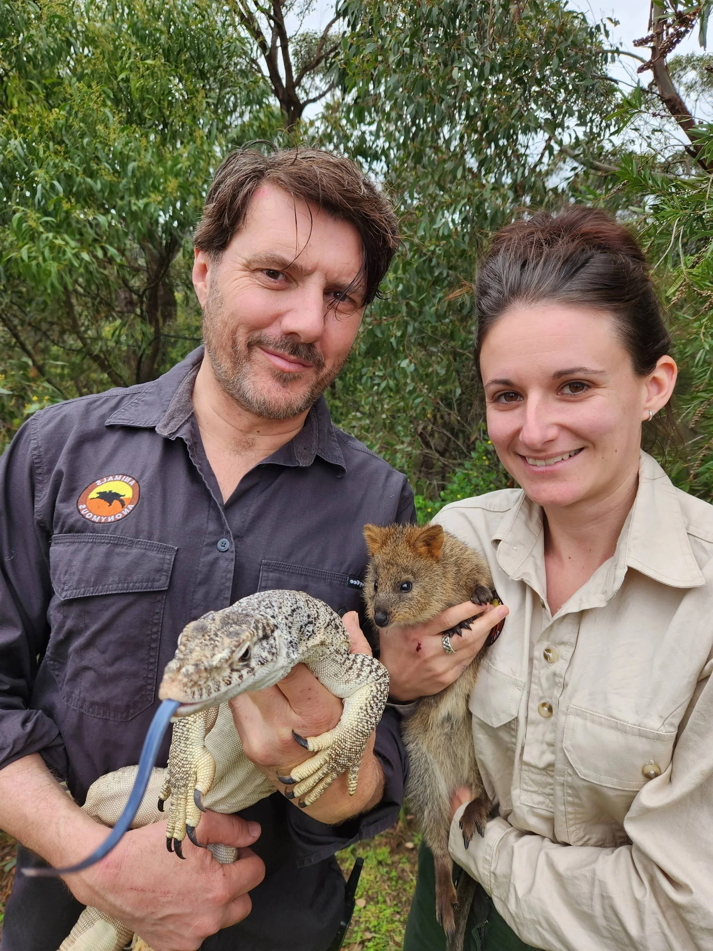 Two people holding a lizard and a quokka in a green outdoor environment.
