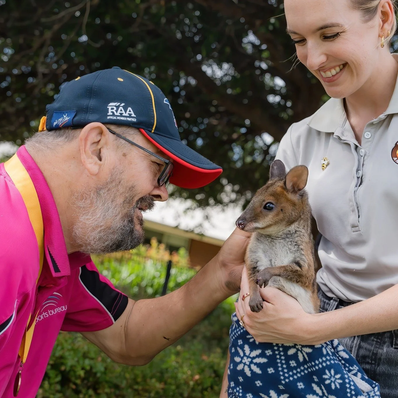 A man with glasses and a beard wearing a pink shirt, and a woman in a light gray polo holding a pademelon smiling.