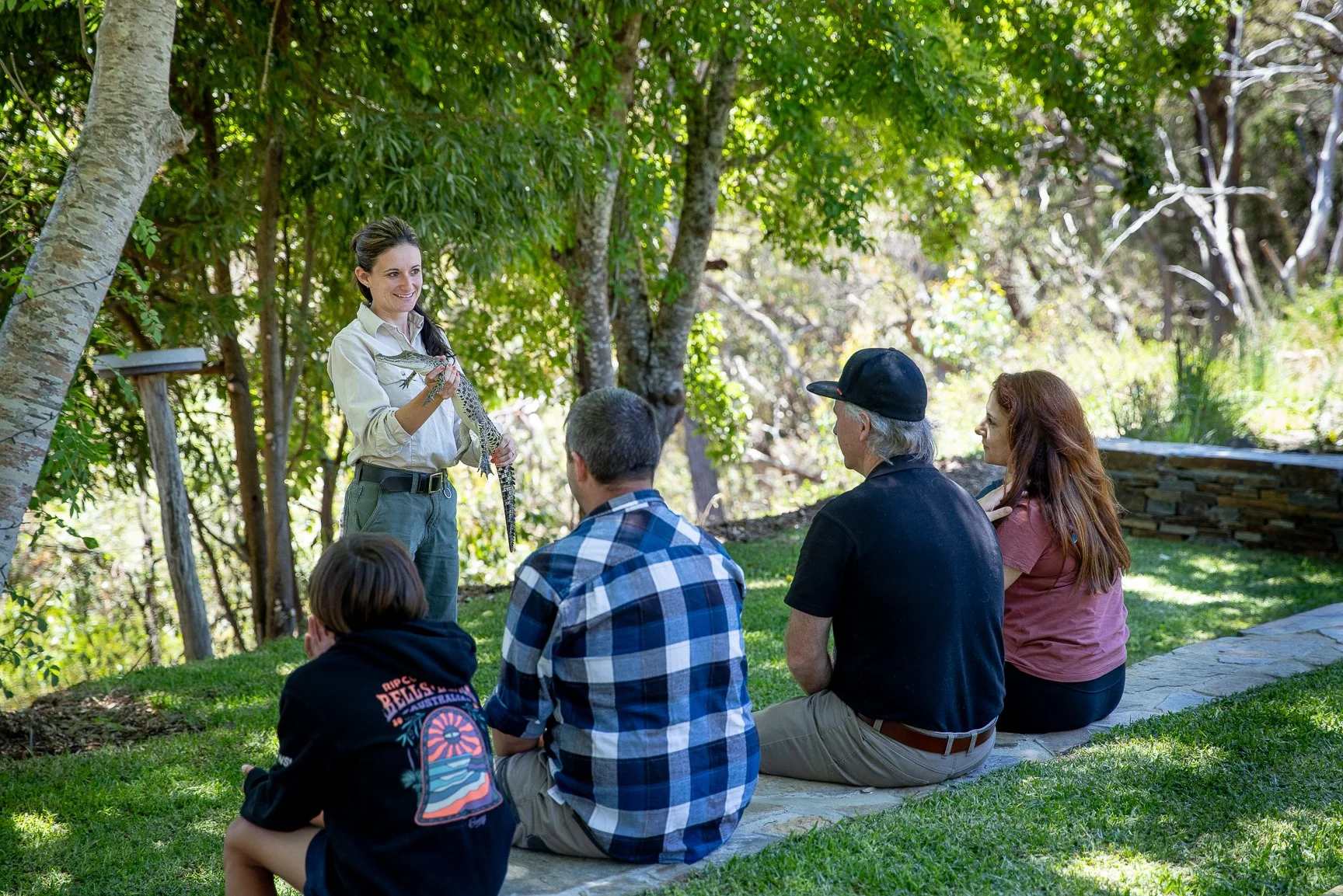 A woman holding a snake and talking to a group of people sitting outdoors on a grassy area with trees in the background.