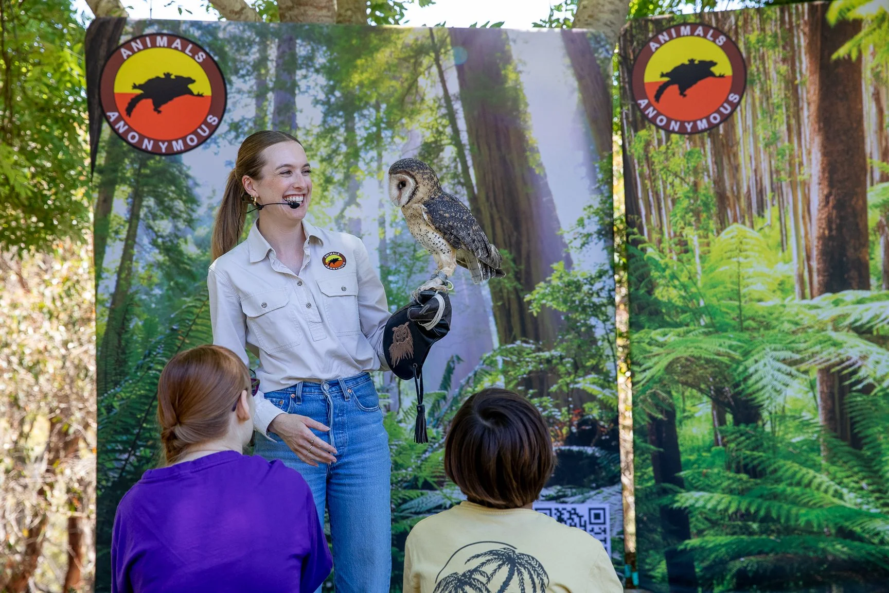 A woman with a microphone headset holding an owl on her gloved hand, speaking to a small audience in front of a backdrop with forest scenery and two circular signs reading "Animals Anonymous".
