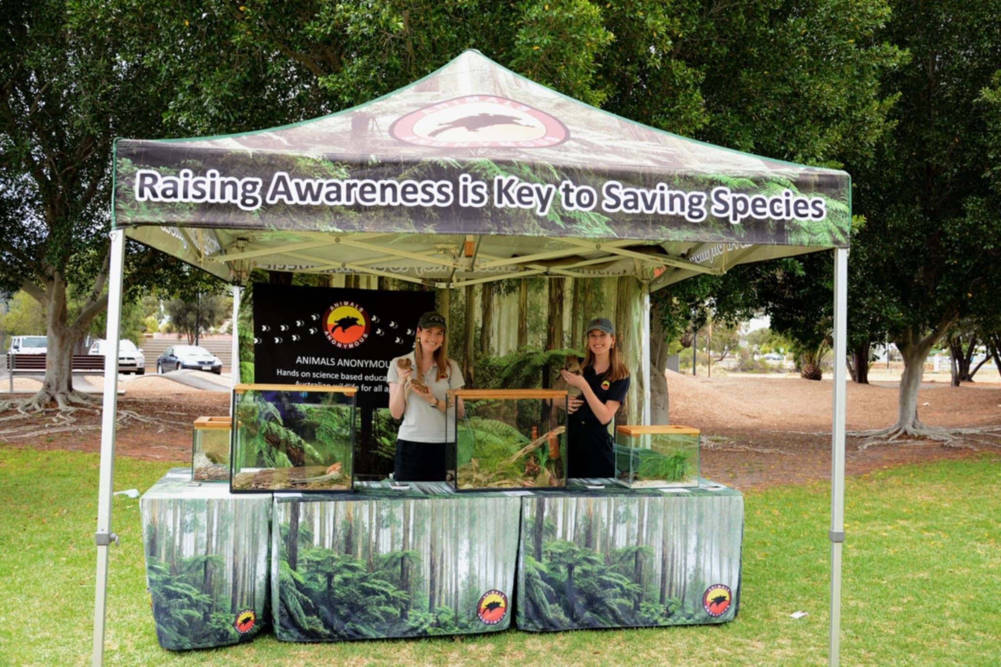 Two women standing inside a display marquee with terrariums, under a canopy that reads 'Raising Awareness is Key to Saving Species.'