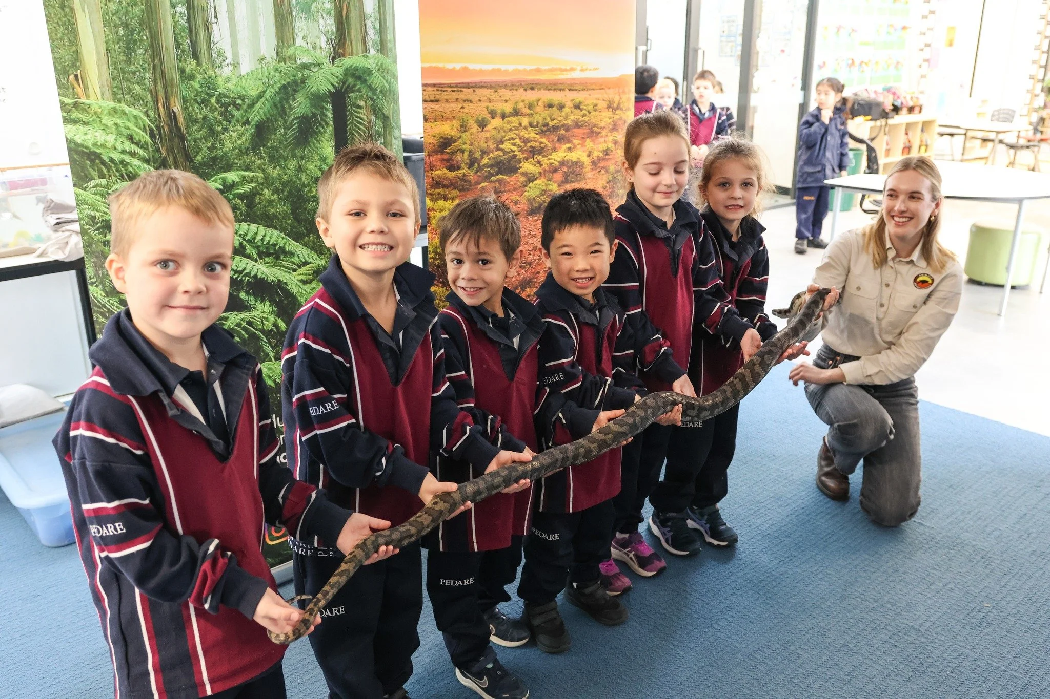 Group of young children holding a large python with a presenter in a classroom with nature-themed backdrop.