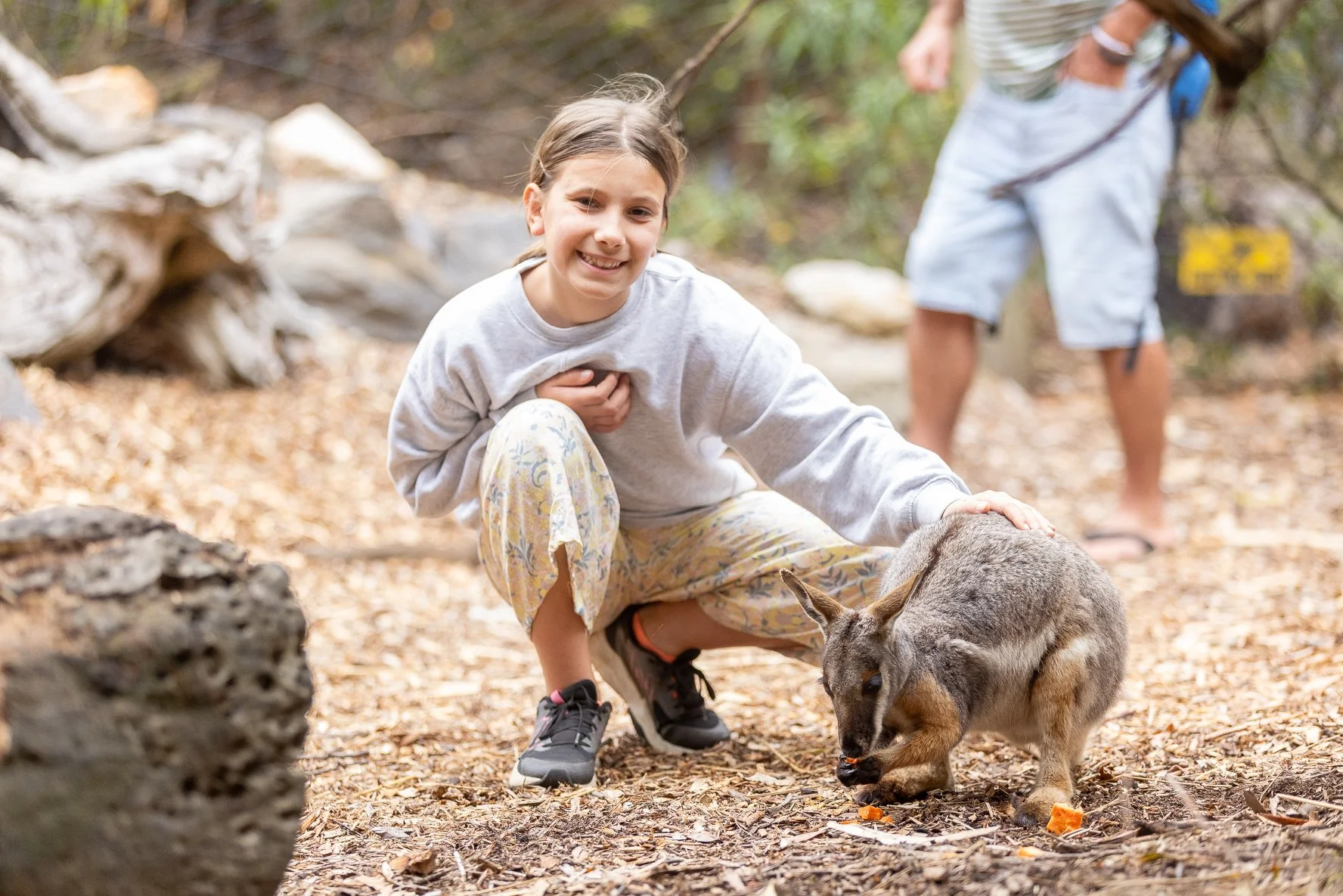 A young girl squatting outdoors, petting a wallaby, on the ground covered with leaves, with another person standing in the background.