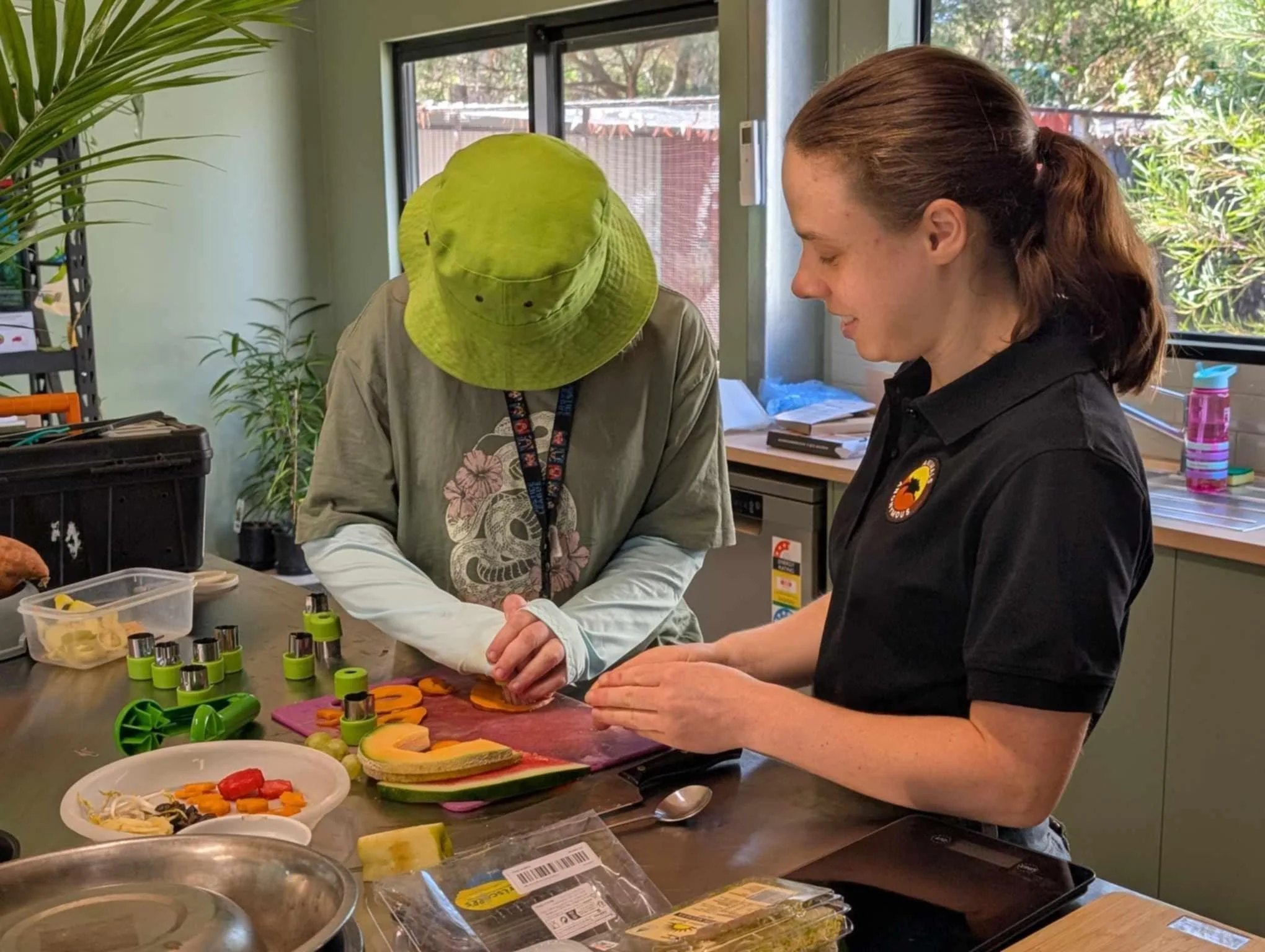 Two women preparing sliced fruits and vegetables on a kitchen counter, with various kitchen tools and containers around them, in a room with large windows.
