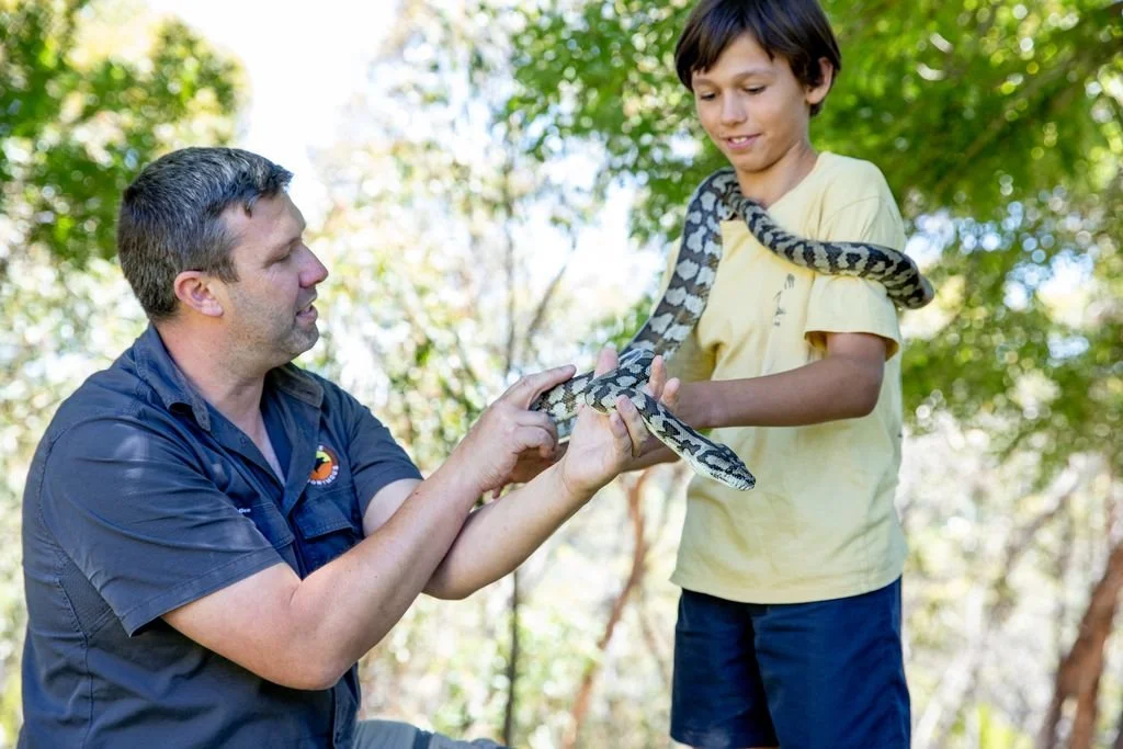 A man and a boy outdoors with trees in the background, examining a large python that the boy is holding.