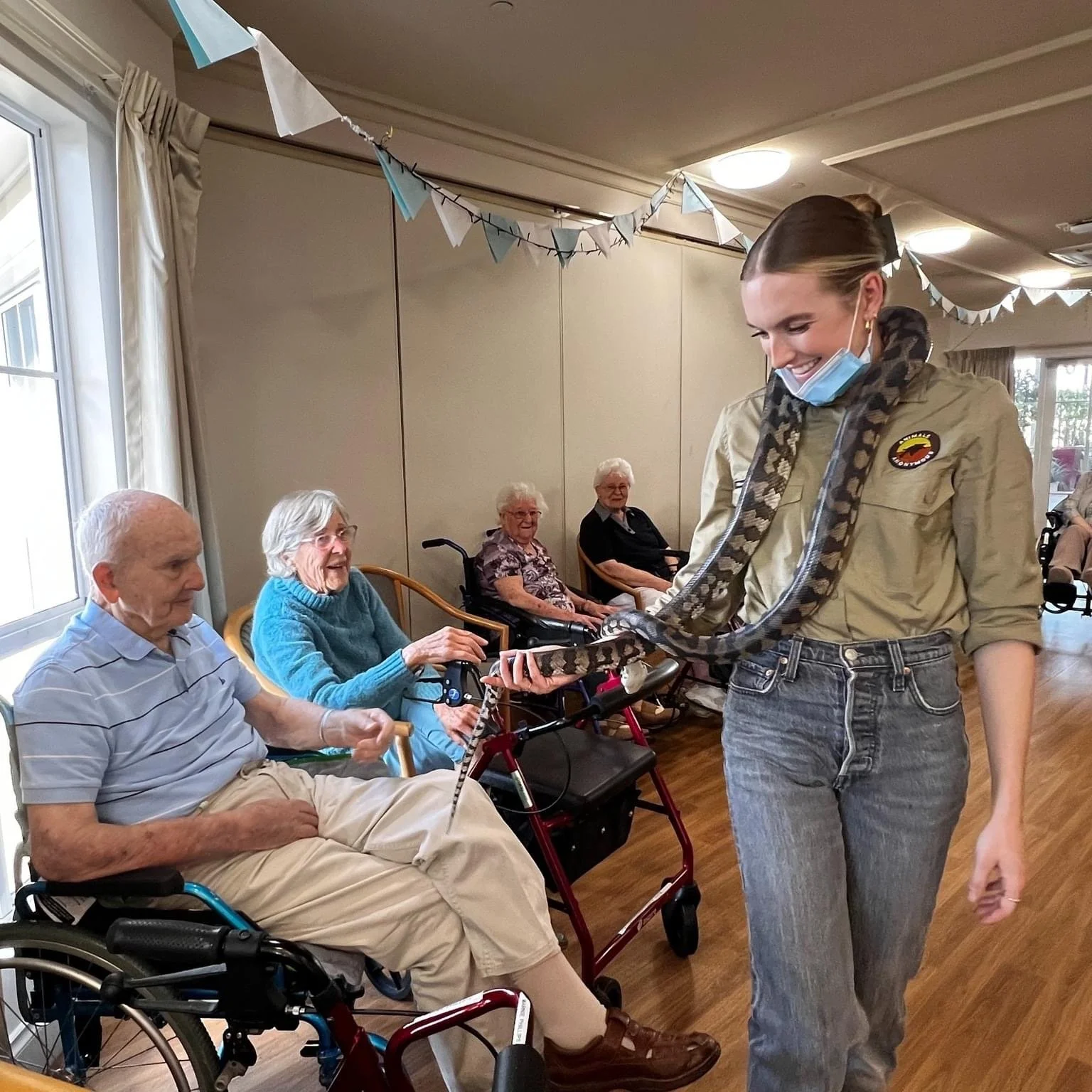 A young woman with a snake draped around her neck and shoulders, smiling at elderly people seated in a room decorated for a celebration or event.