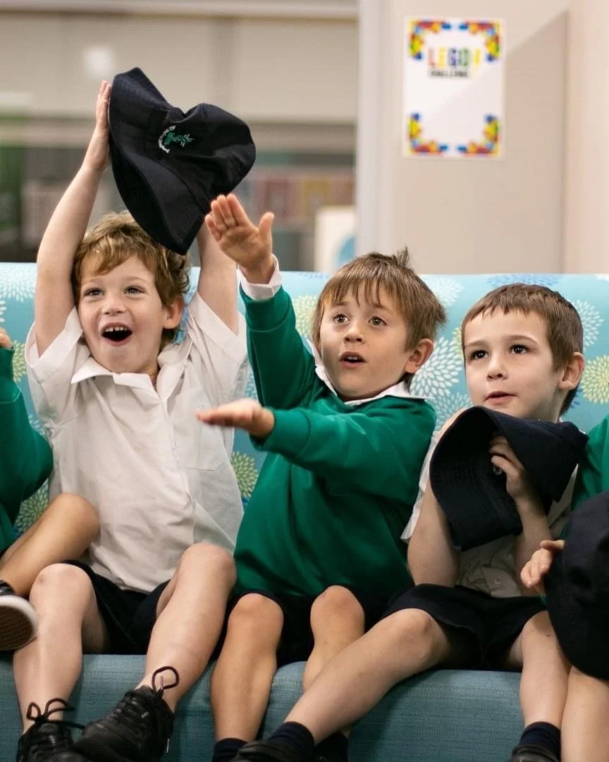 Three young boys in school uniforms sitting on a couch in a classroom, with one boy raising his hat excitedly.
