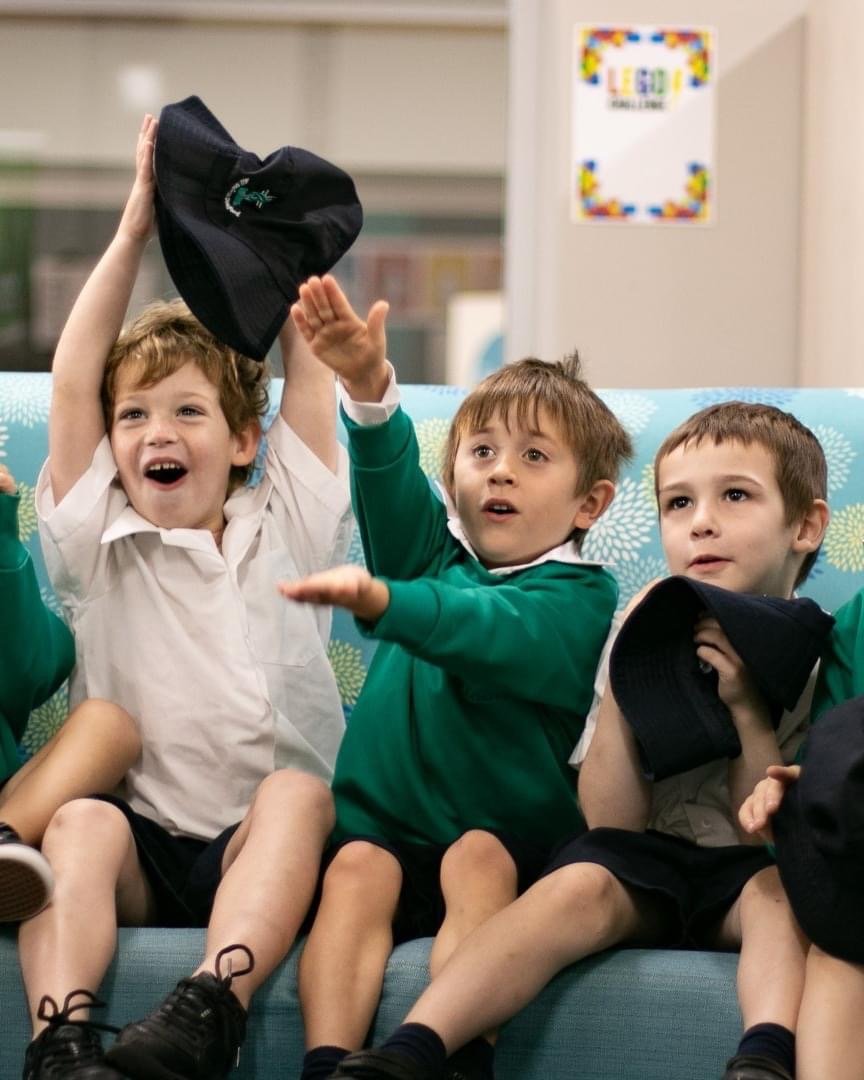 Three young boys in school uniforms sitting on a couch in a classroom, with one boy raising his hat excitedly.