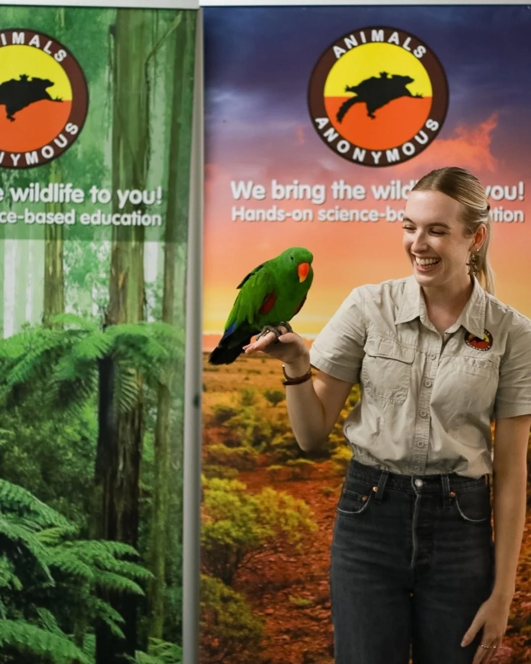 A woman holding an eclectus parrot on her hand, standing in front of banners with wildlife and nature themes from Animals Anonymous.