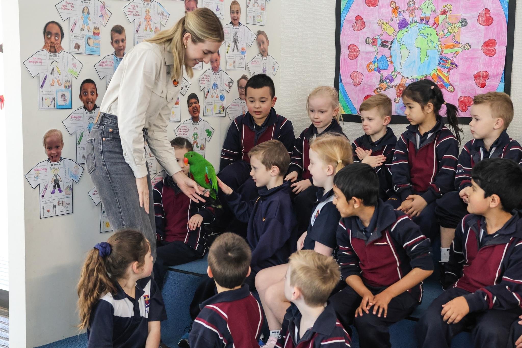 Classroom scene with a woman and children. The woman is showing a green parrot to the children, who are sitting on the floor. The background has colorful artwork and posters, including children's drawings of themselves and a large poster with children holding hands around a globe.