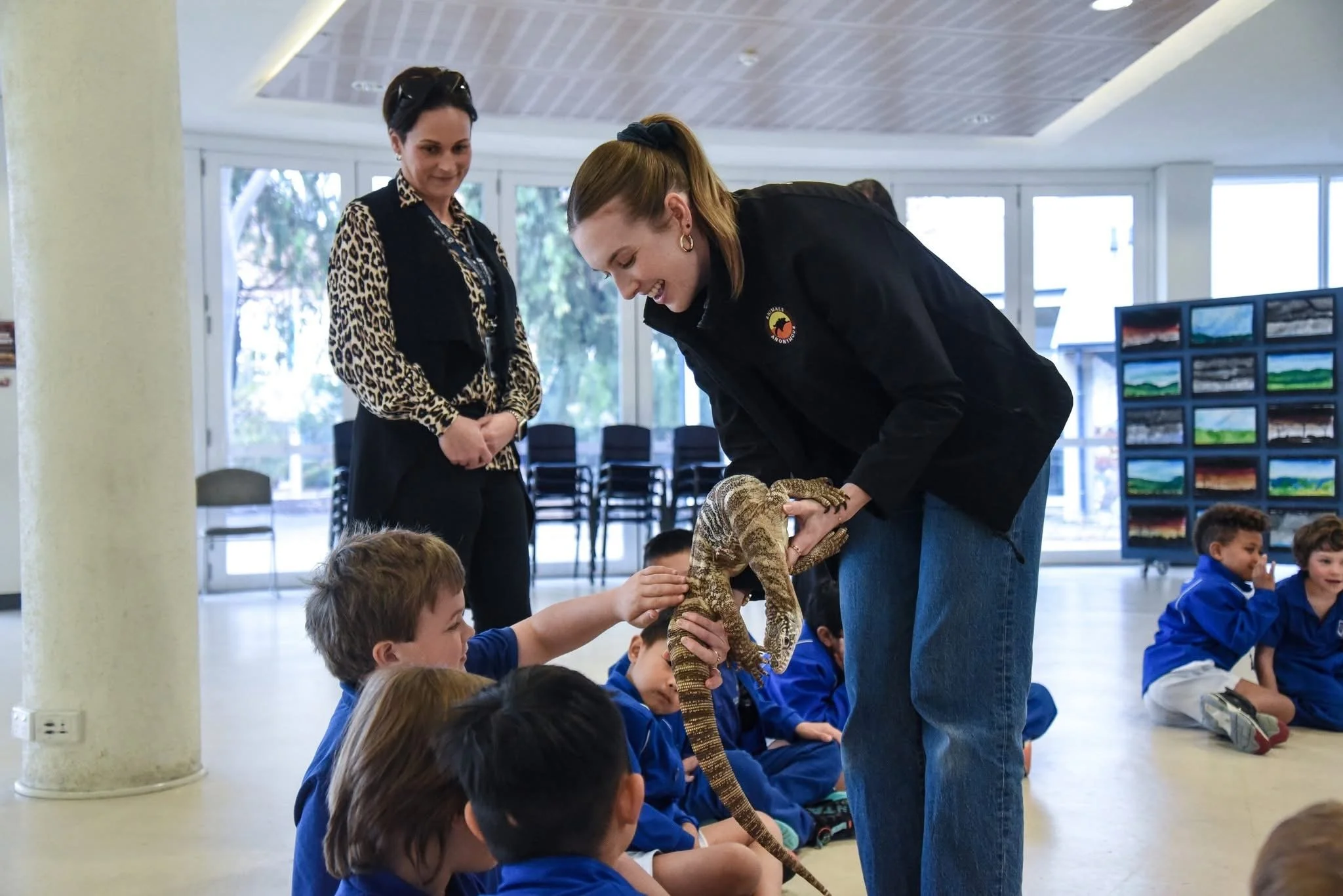 Young children sitting on the floor in a classroom, patting a goanna from an adult woman during an educational presentation. Other women are present, and there are display boards with landscape images in the background.
