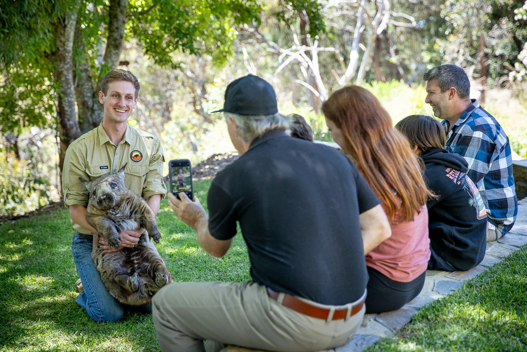 A young man in a tan uniform kneels on the grass holding a young wombat, smiling, as a group of people sit on a stone path watching and taking photos in a forested park.