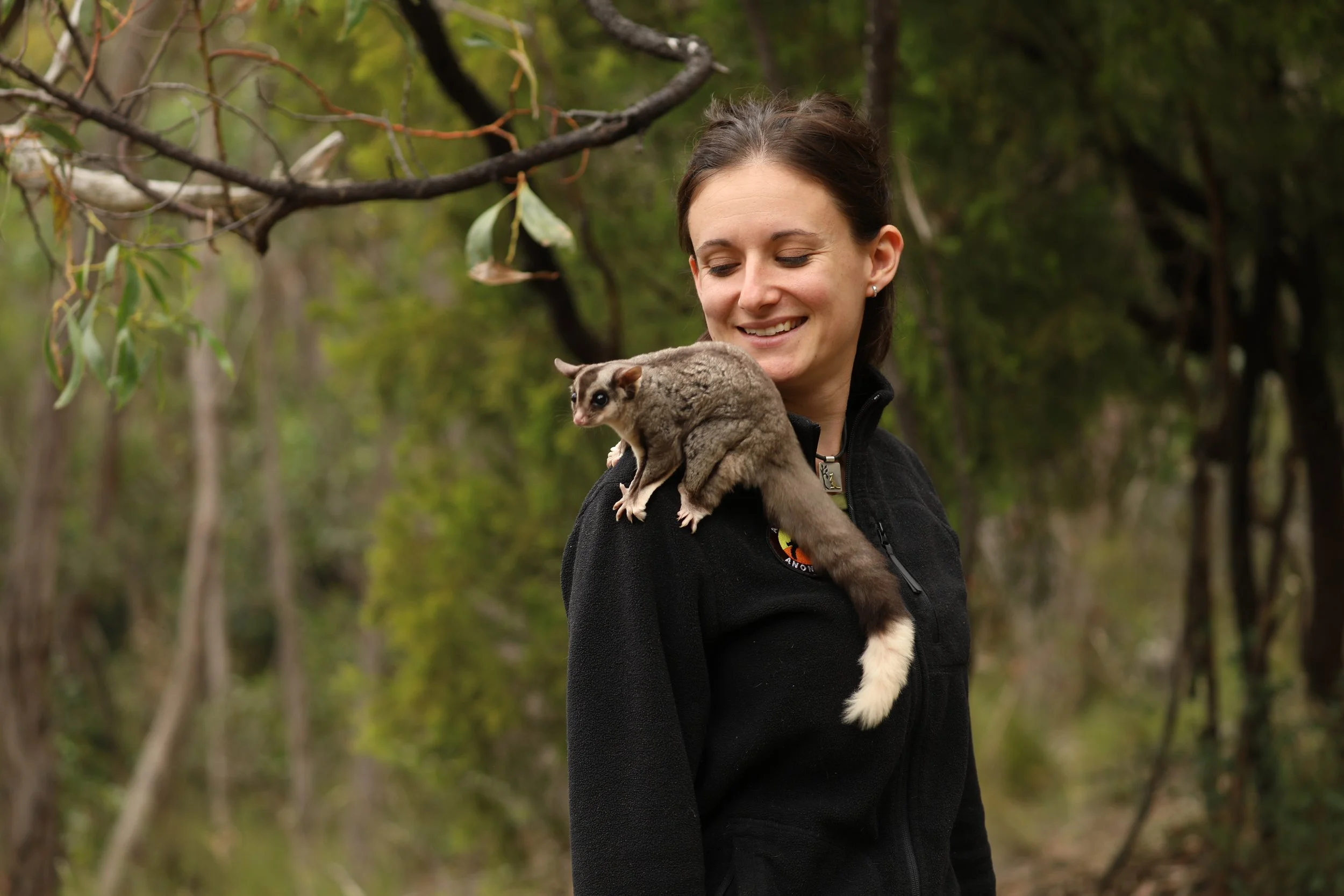 A woman wearing a black jacket standing in a forest, smiling as a squirrel glider rests on her shoulder.