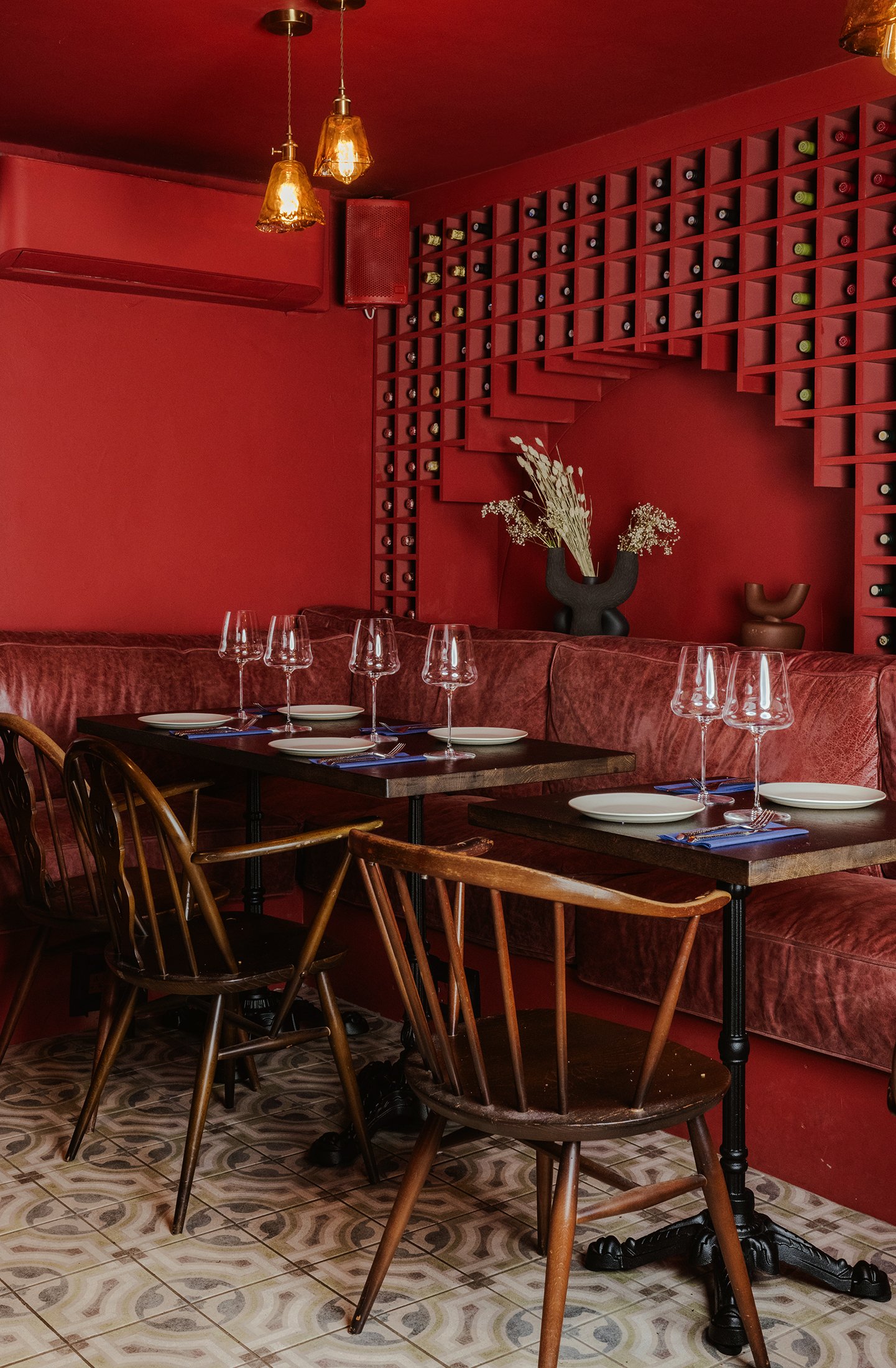 A cozy restaurant interior with red walls and booth seating. There are wine bottles arranged on the wall in a decorative pattern. The tables are set with wine glasses, plates, and utensils, and wooden chairs are placed around them. Two pendant lights