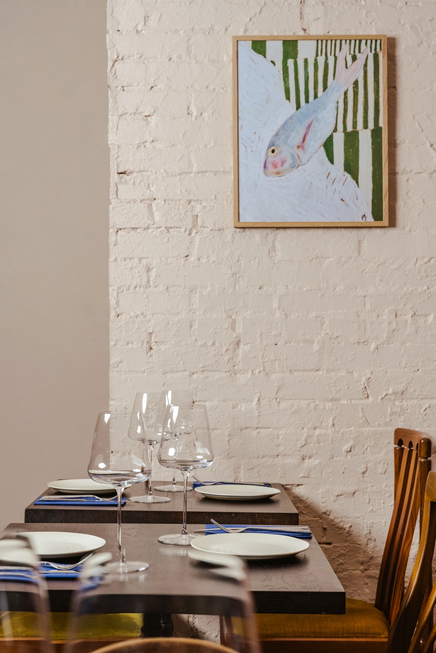 A dining area with a wooden table set for a meal, including white plates, blue napkins, and wine glasses. A painting of a fish on a white brick wall is in the background.