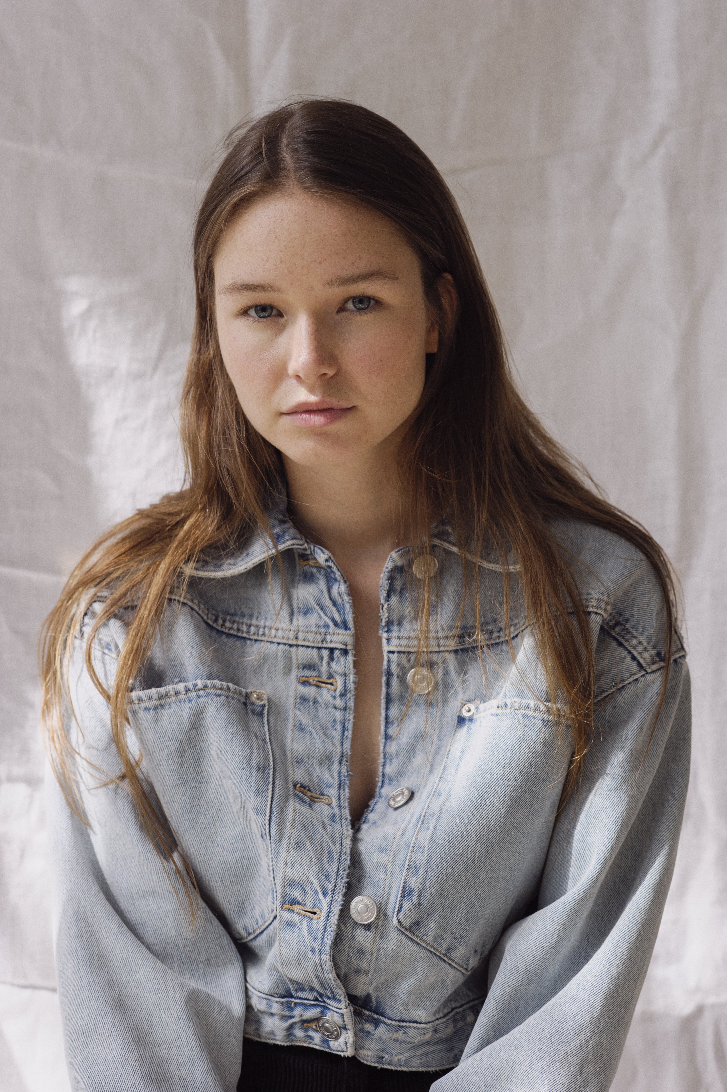 A young woman with long brown hair, blue eyes, and freckles, wearing a light denim jacket, posing in front of a neutral fabric backdrop.