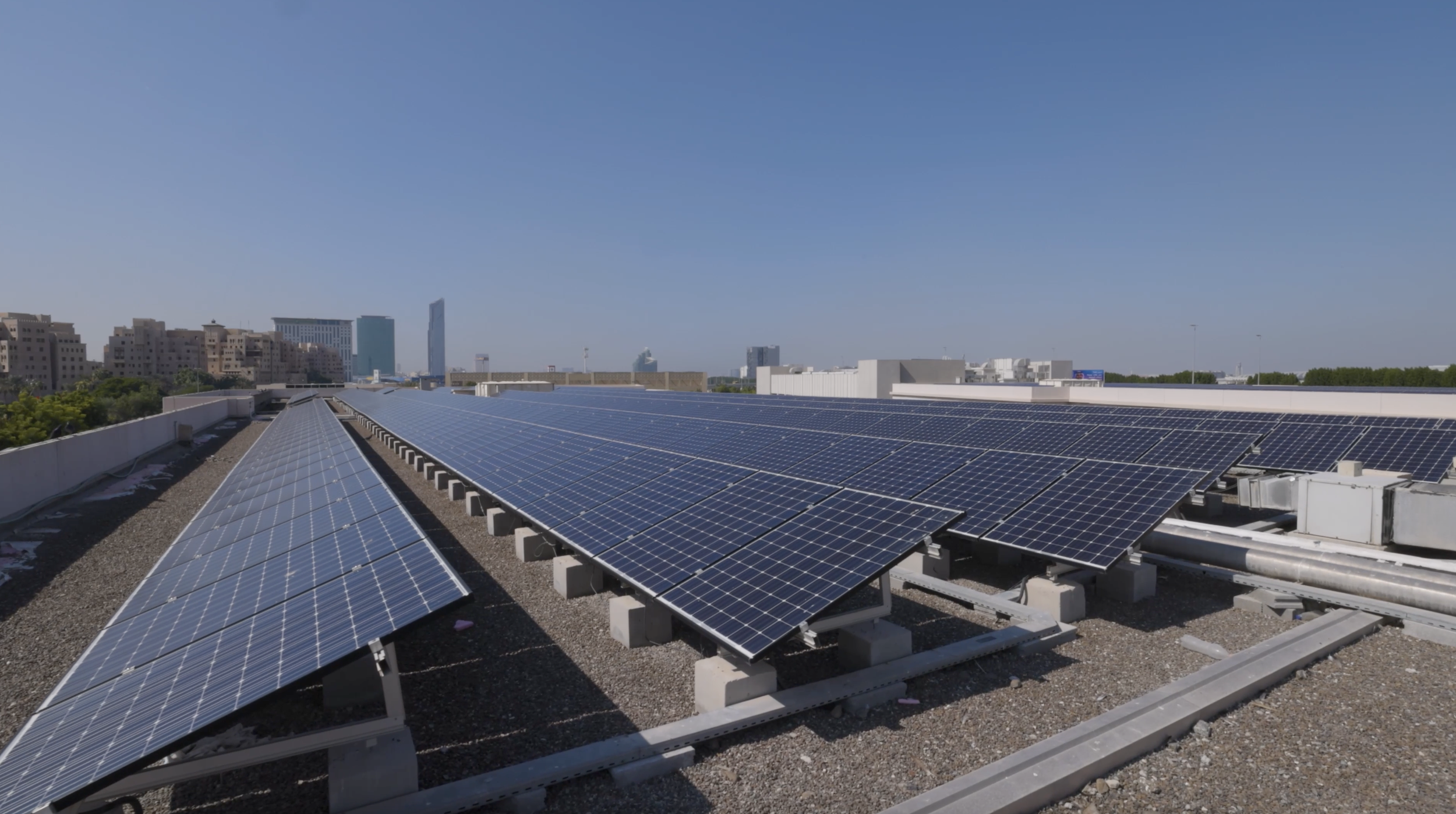 A rooftop covered with solar panels against a clear blue sky, with a city skyline featuring tall buildings in the background.