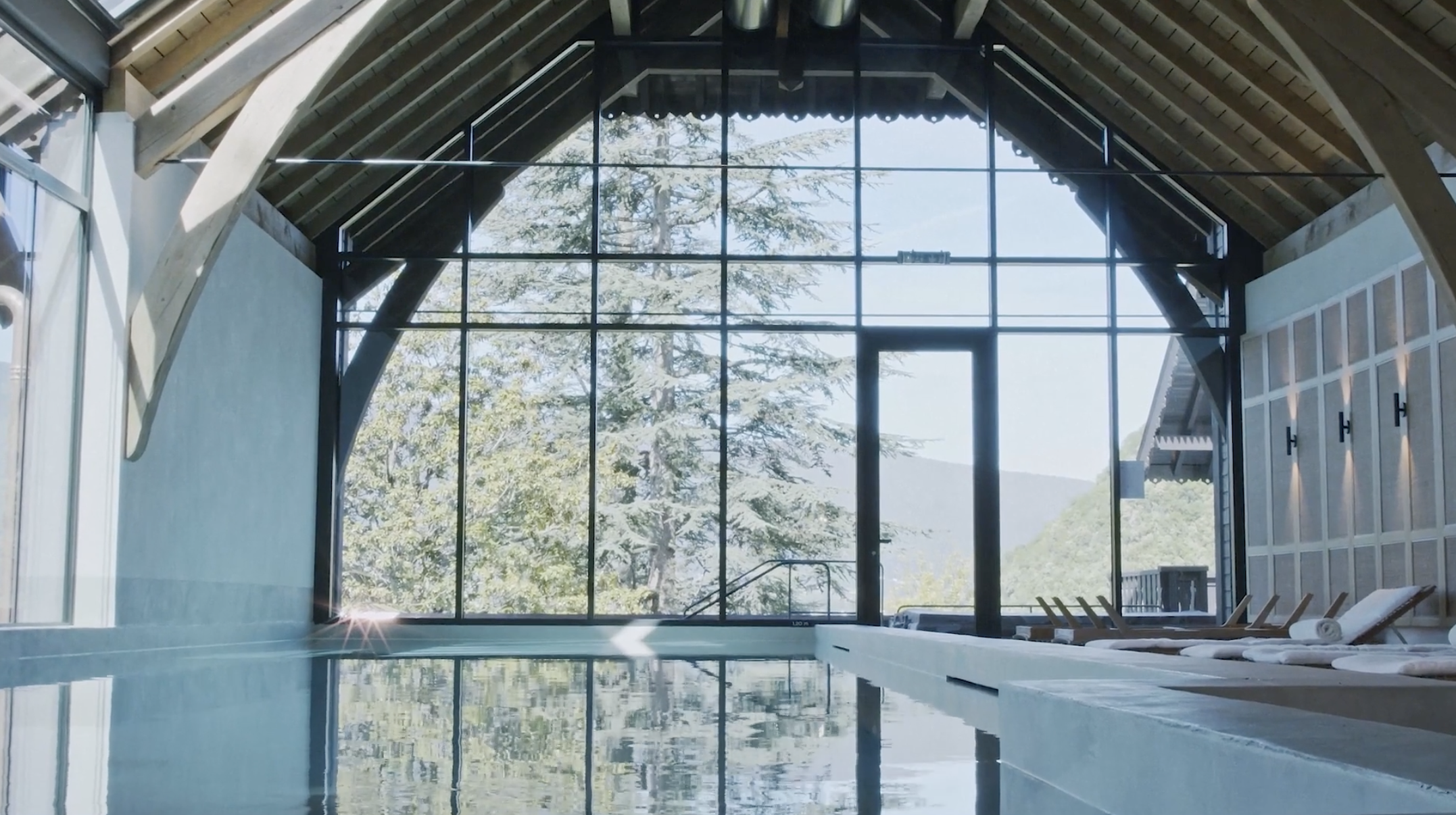 Indoor swimming pool with large glass windows showing trees and mountains outside, wooden beams supporting the ceiling, and lounge chairs along the poolside.