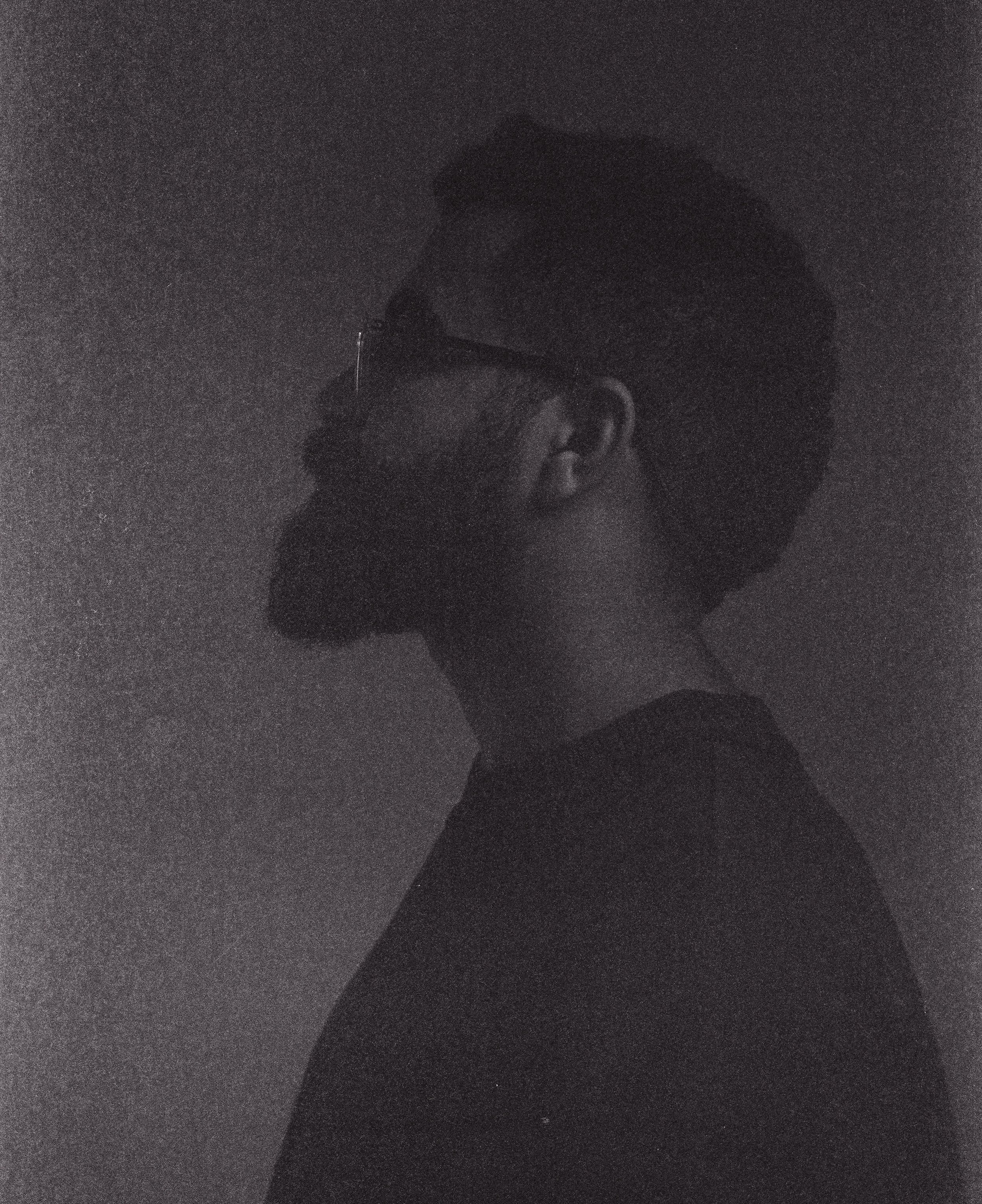 A black and white side profile of a man with a beard, glasses, and styled hair, against a plain background.