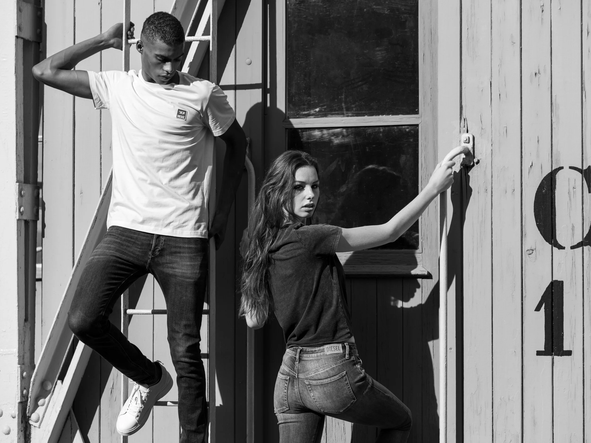 A black and white photo of a young man and woman posing outside a wooden building. The man, on a ladder, rests his arm on his head, while the woman, leaning against the wall, holds a handle.