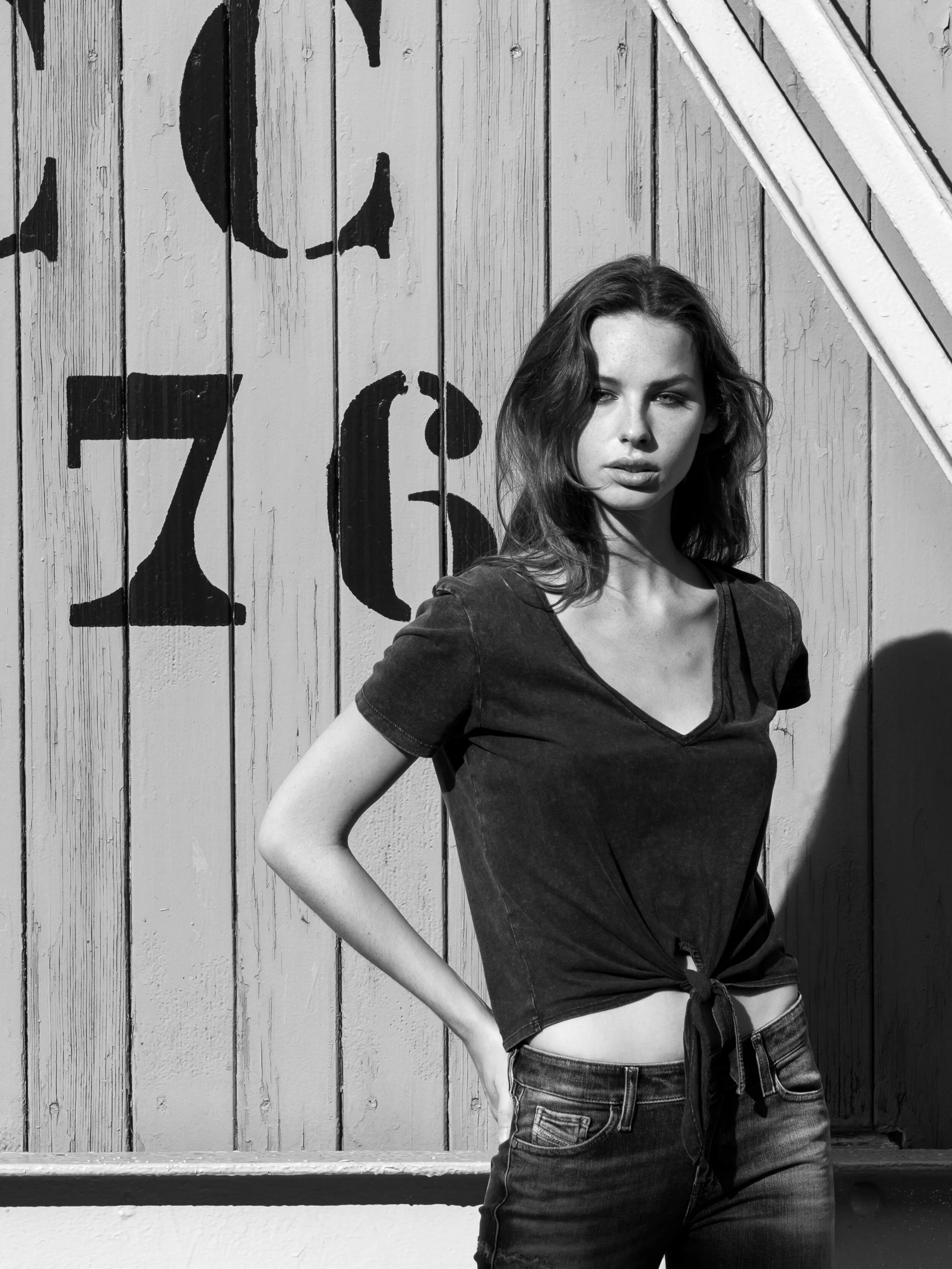 A young woman with shoulder-length hair stands against a wooden wall with painted numbers and letters, wearing a tied-up t-shirt and jeans, casting a shadow on the wall.