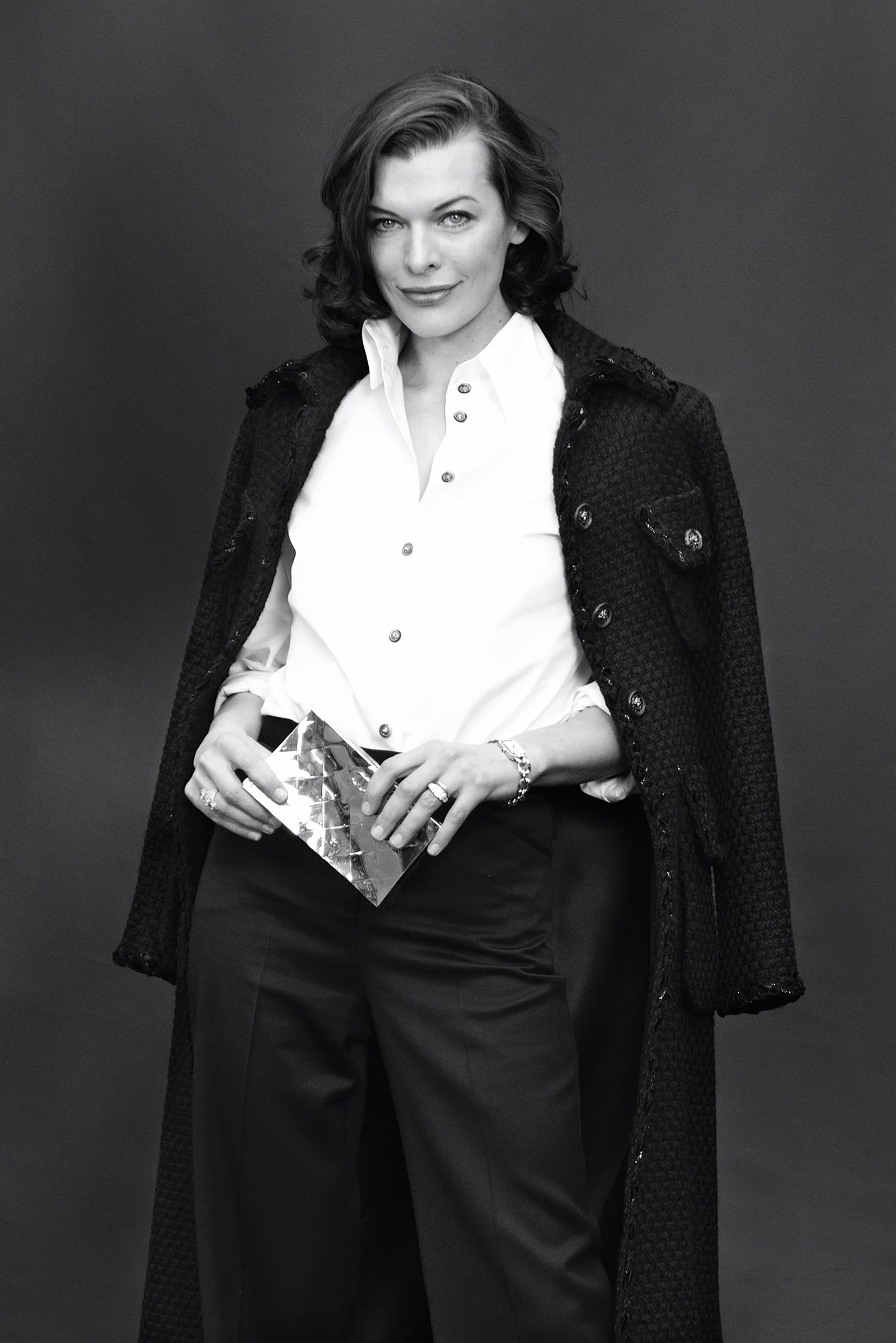 Black and white photograph of a woman with medium-length wavy hair, wearing a white button-up shirt, dark skirt, and a textured coat draped over her shoulders. She is holding a shiny, reflective clutch purse with both hands, adorned with jewelry including rings, a bracelet, and a watch. The background is plain and dark.