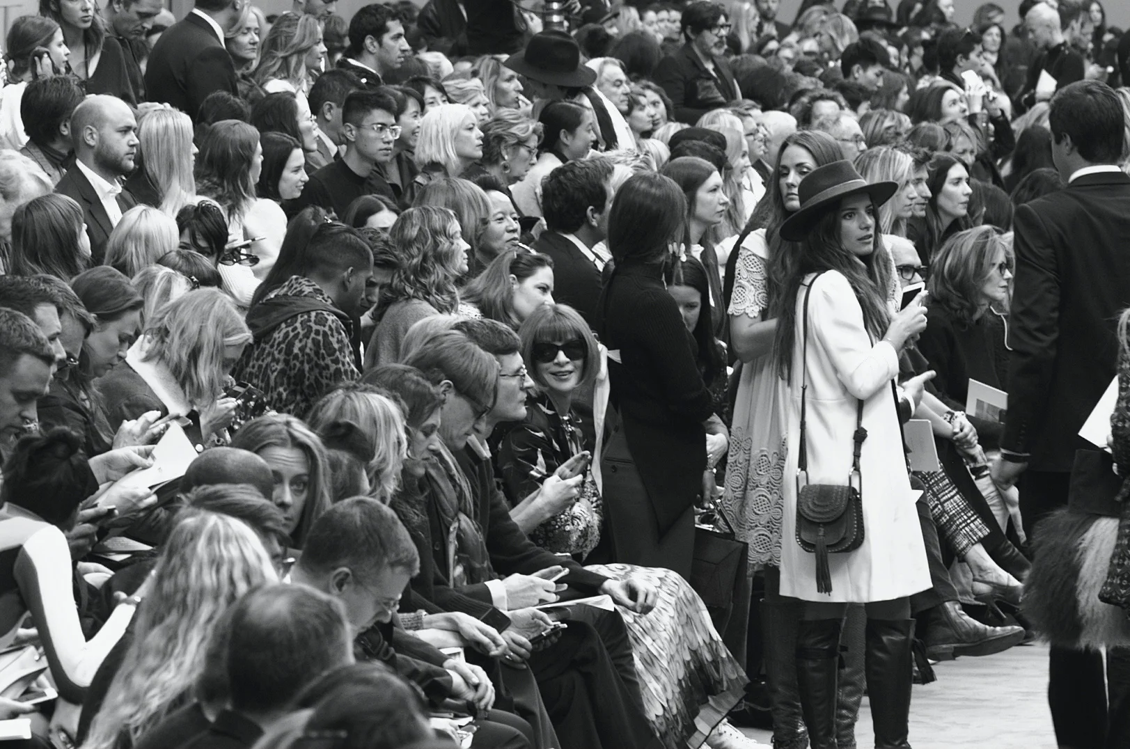 Crowd of people attending an event, with some looking at their phones and others watching or waiting.