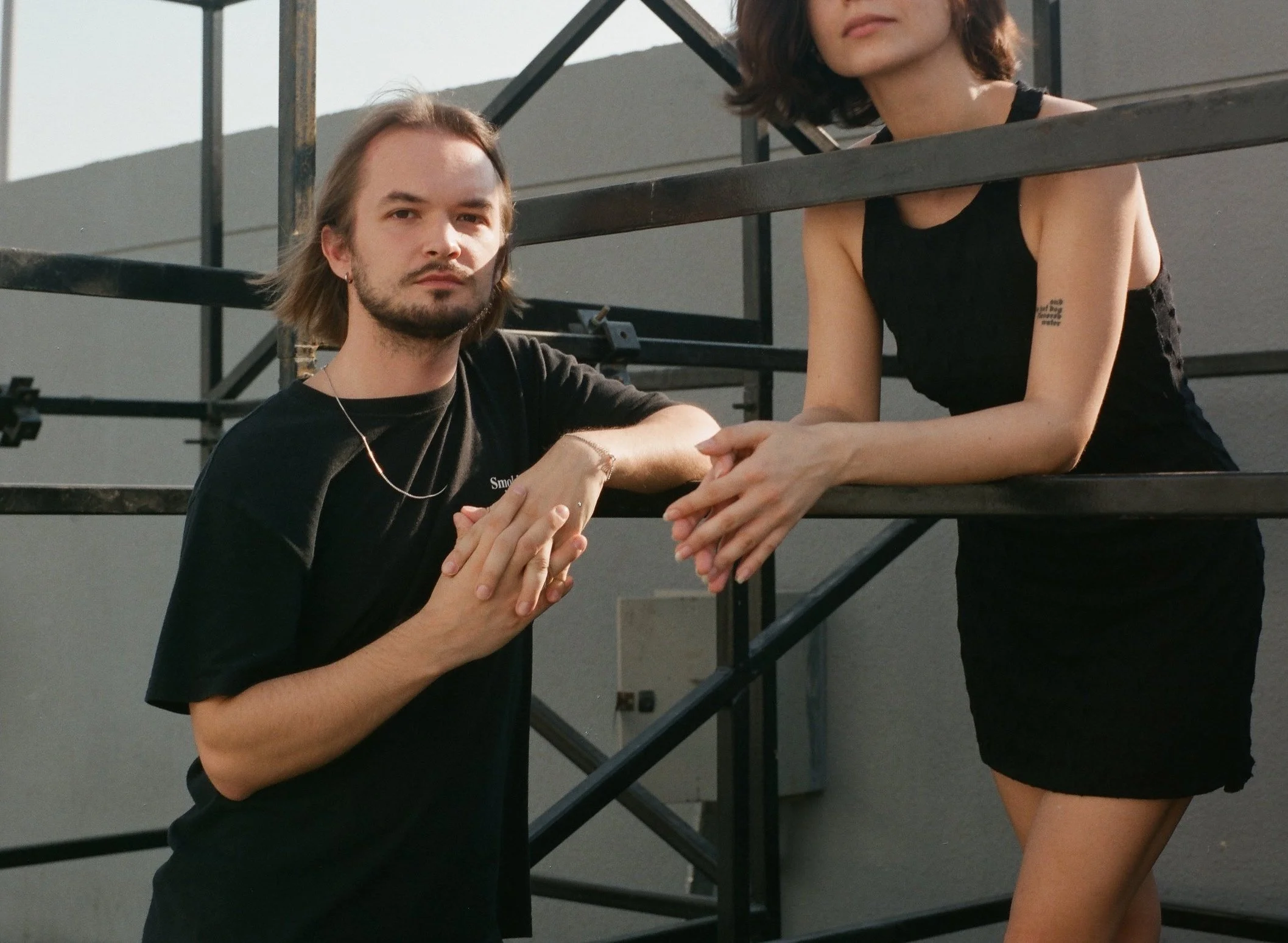 A young man with shoulder-length hair, a beard, and wearing a black t-shirt and a silver necklace, standing outdoors in front of a black metal staircase, with a woman in a black sleeveless dress, with tattoo on her left upper arm, leaning on the rail