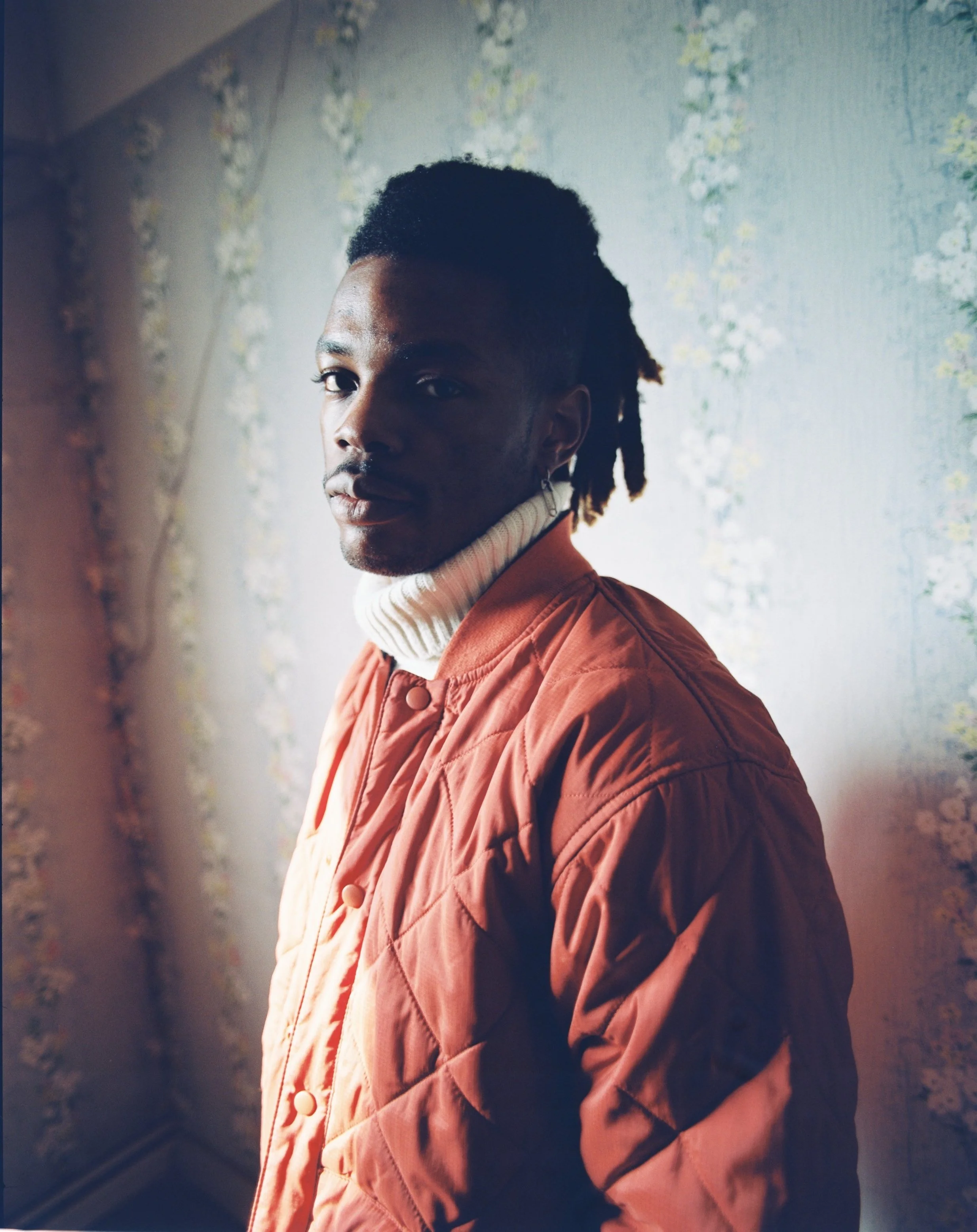 Portrait of a young man with dark skin, wearing a white turtleneck and an orange quilted jacket, standing in front of a floral wallpaper background, looking at the camera.