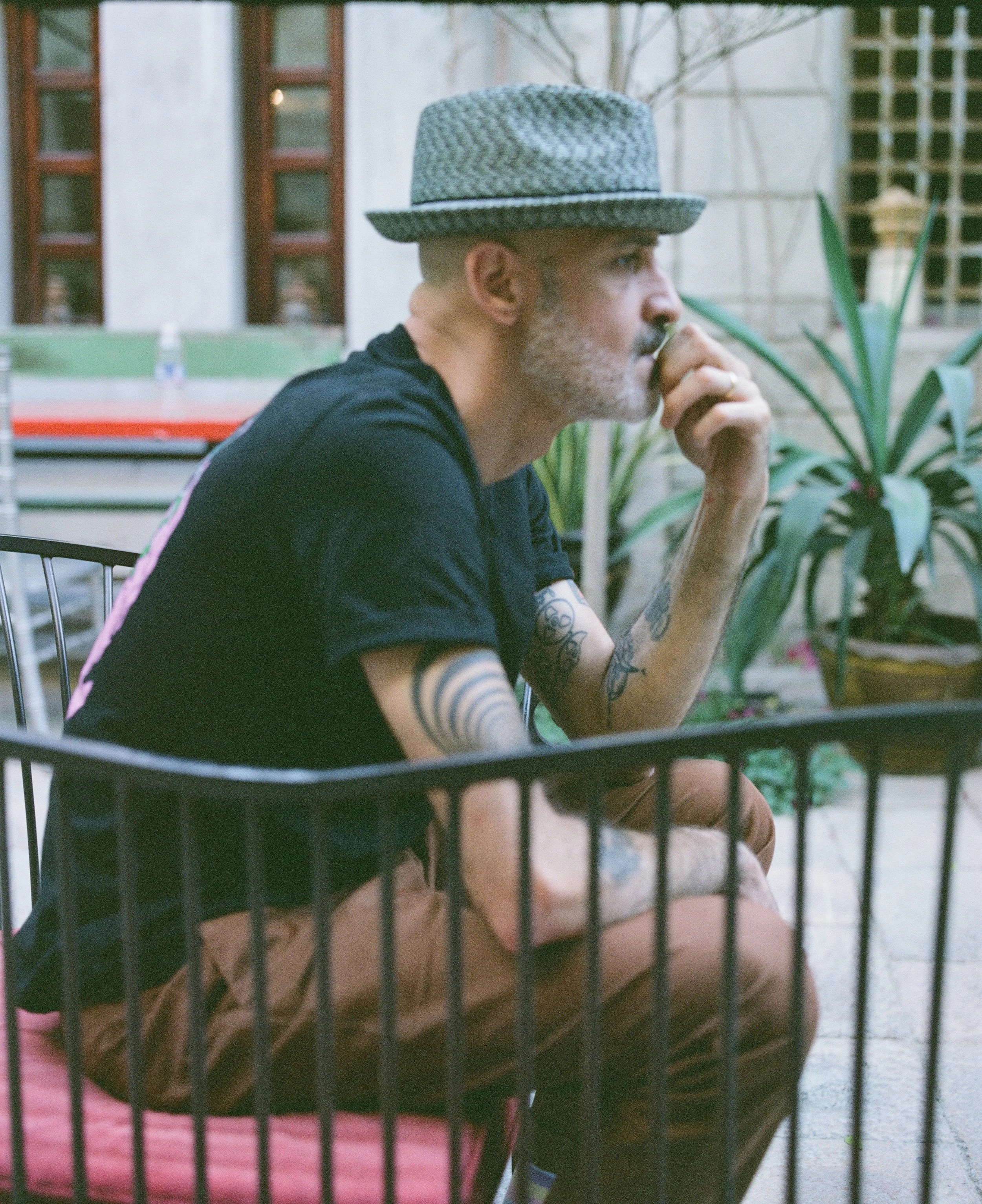 A man with tattoos, wearing a patterned hat and black t-shirt, sitting in a card with a pink cushion outside, surrounded by plants and a building in the background, resting his chin on his hand.