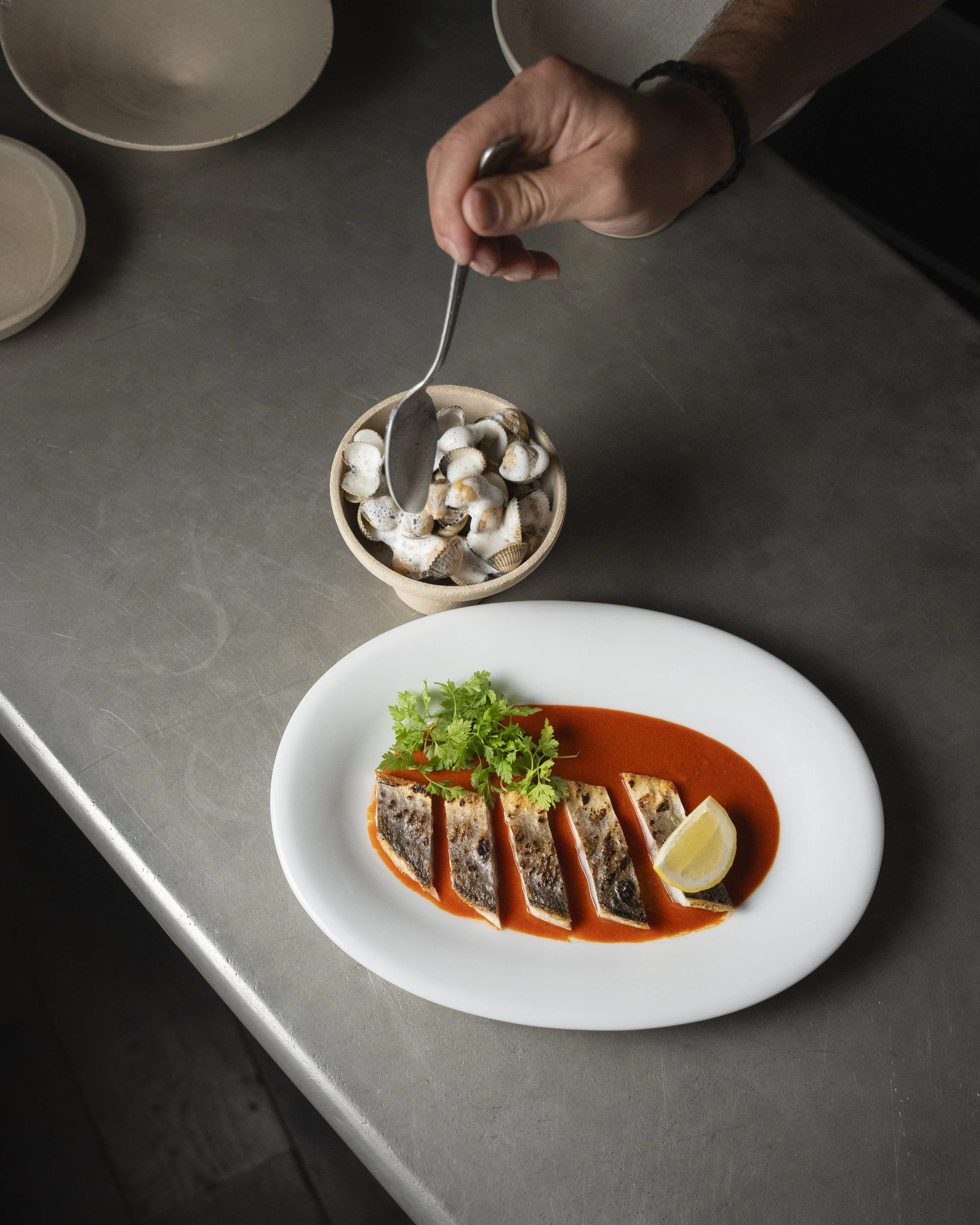 A plate of sliced fish with tomato sauce, a lemon wedge, and fresh herbs, with a bowl of shellfish and a hand holding a spoon above it.