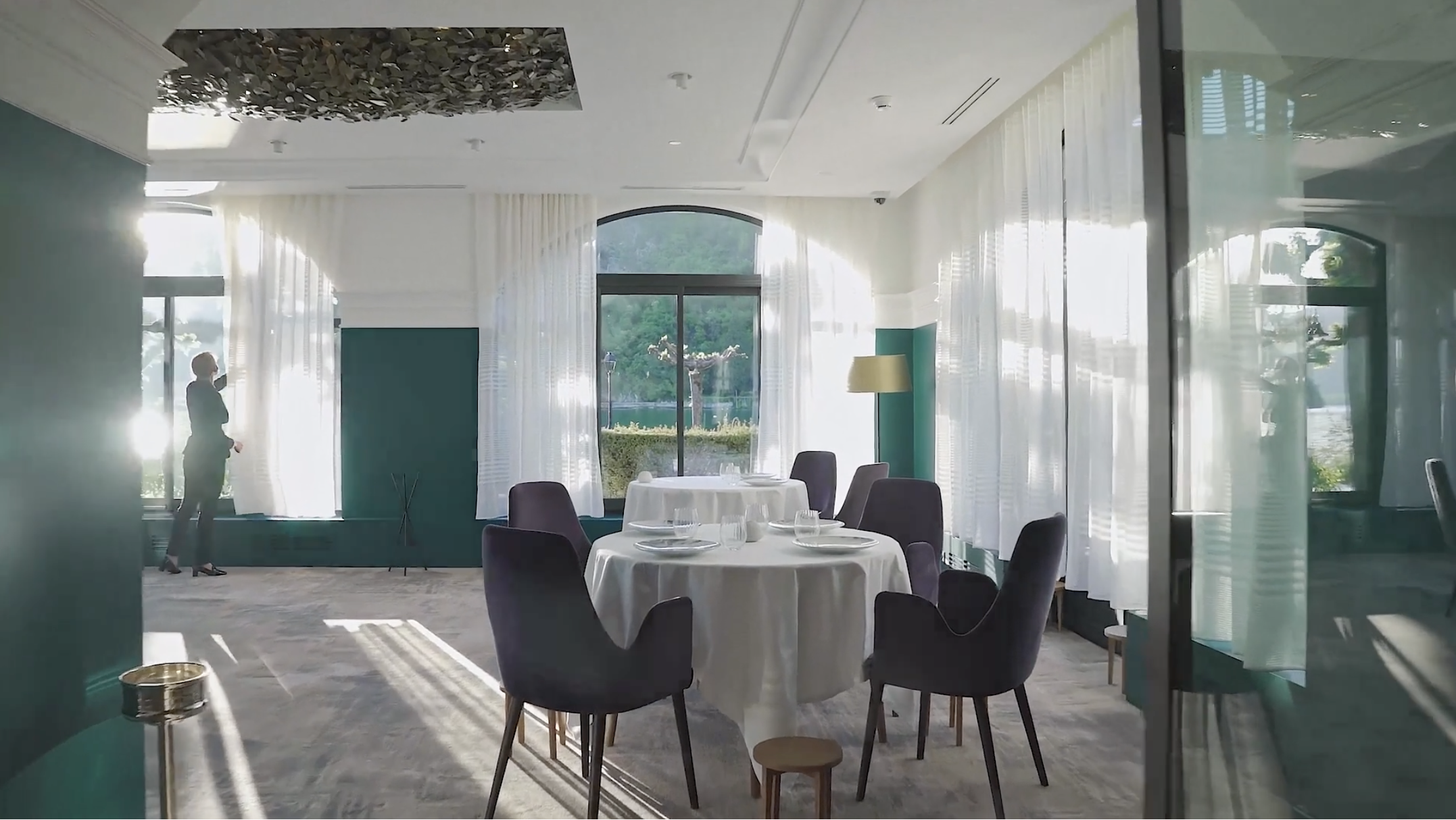 Elegant dining room with round tables covered with white tablecloths, set with glassware and plates, surrounded by dark upholstered chairs. Tall windows with sheer white curtains let in natural light, revealing a green outdoor landscape. A standing l