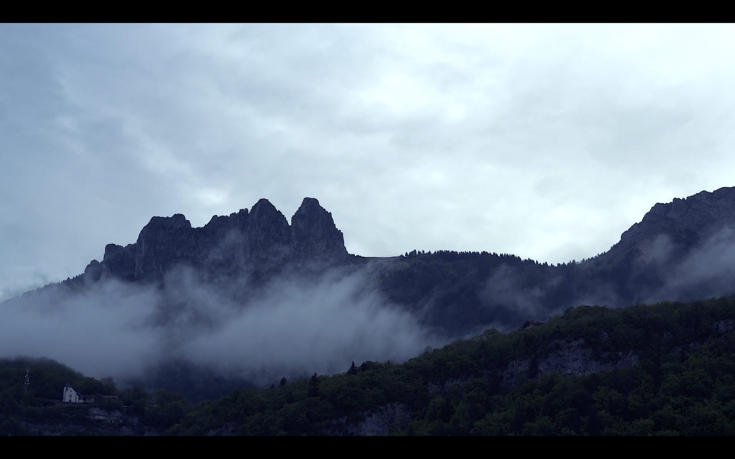 Mountain range with clouds and fog, small structures at the base of the mountains, and a cloudy sky.