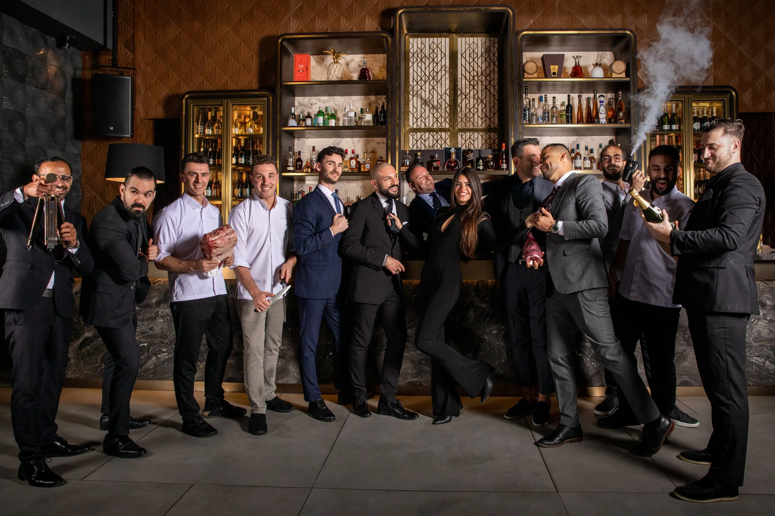 Group of people at a bar, dressed in formal and chef attire, celebrating with various props and drinks, with a well-stocked bar in the background.
