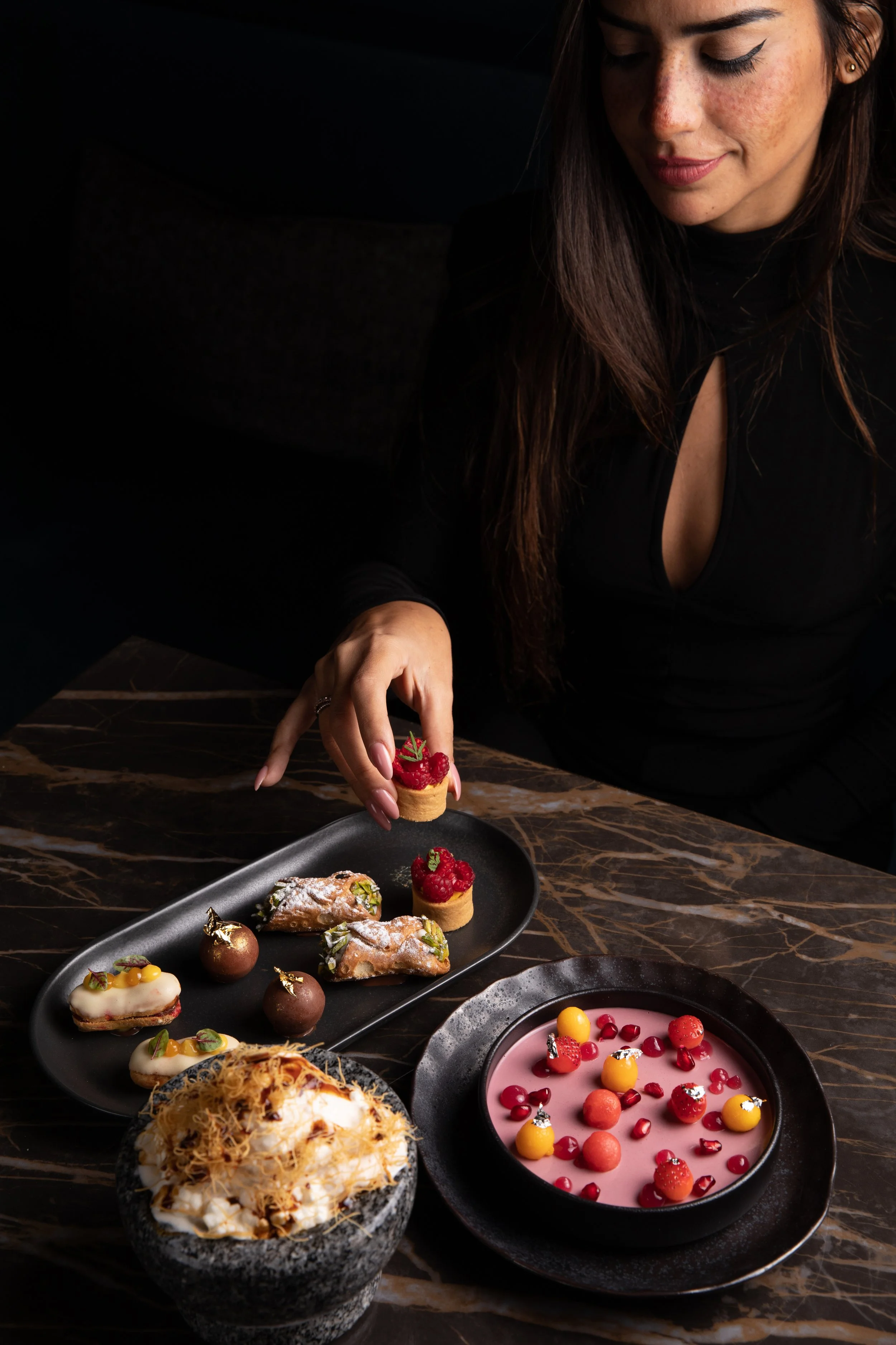 A woman in black clothing with makeup, sitting at a dark marble table, is reaching to pick up a small tart topped with raspberries and a green garnish. The table has a black plate with assorted desserts, including small pastry puffs with powdered sug