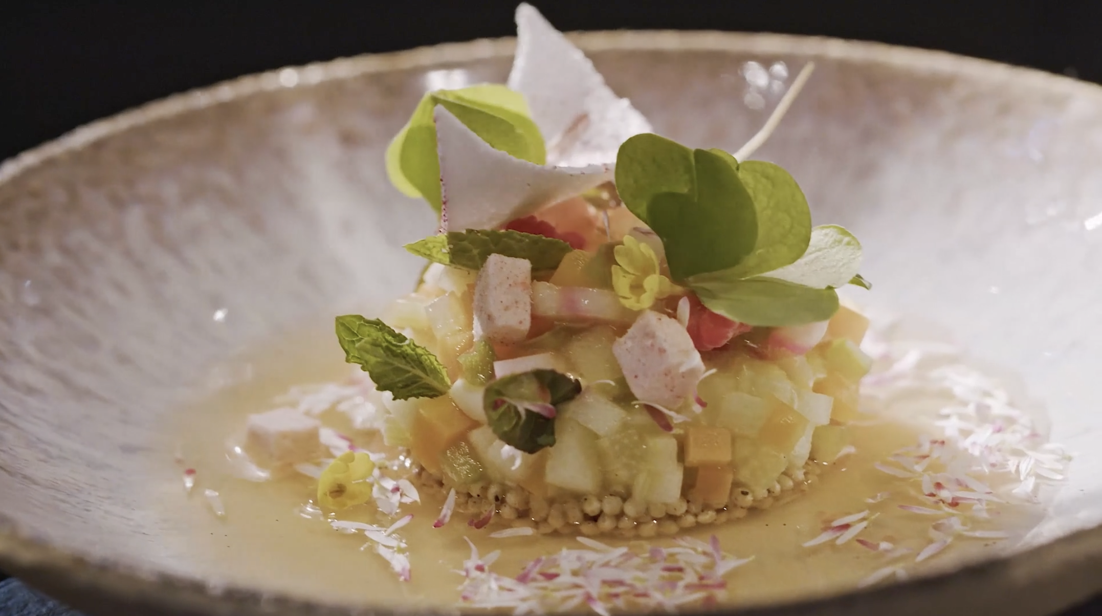 A finely plated dish with chopped vegetables, garnished with fresh herbs and edible flowers, served on a textured ceramic plate.