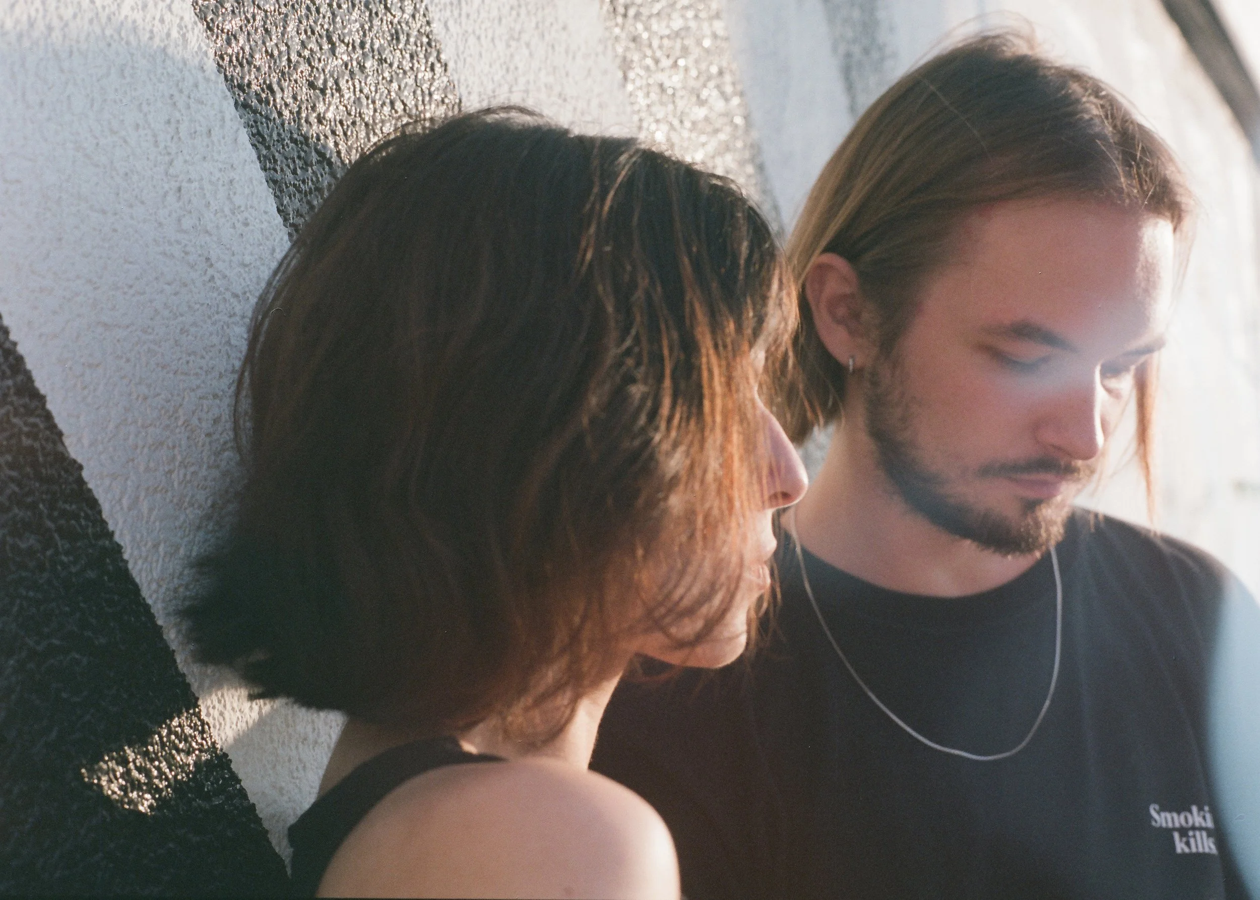A young woman and man sitting close together against a textured wall, their heads bowed and eyes closed, sharing an intimate moment.