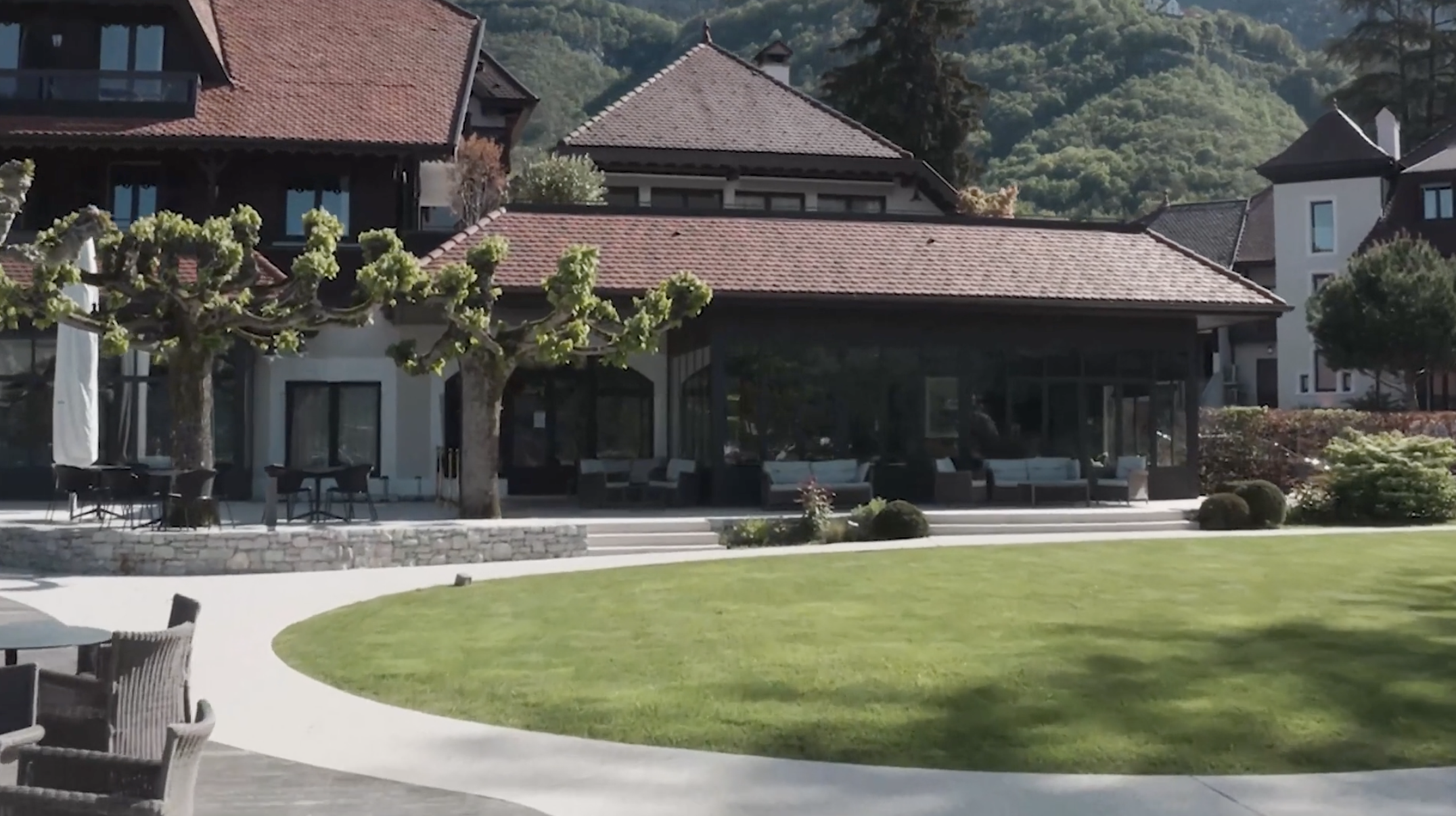 A luxurious house with a red-tiled roof and large glass windows, surrounded by a well-maintained lawn and trees with pruned branches, set against a backdrop of hills.