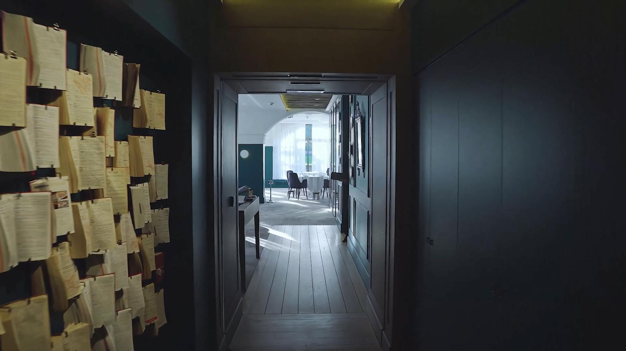 A narrow hallway leading to a bright room with round table and chairs, illuminated by natural light from large windows, with books on the left wall.